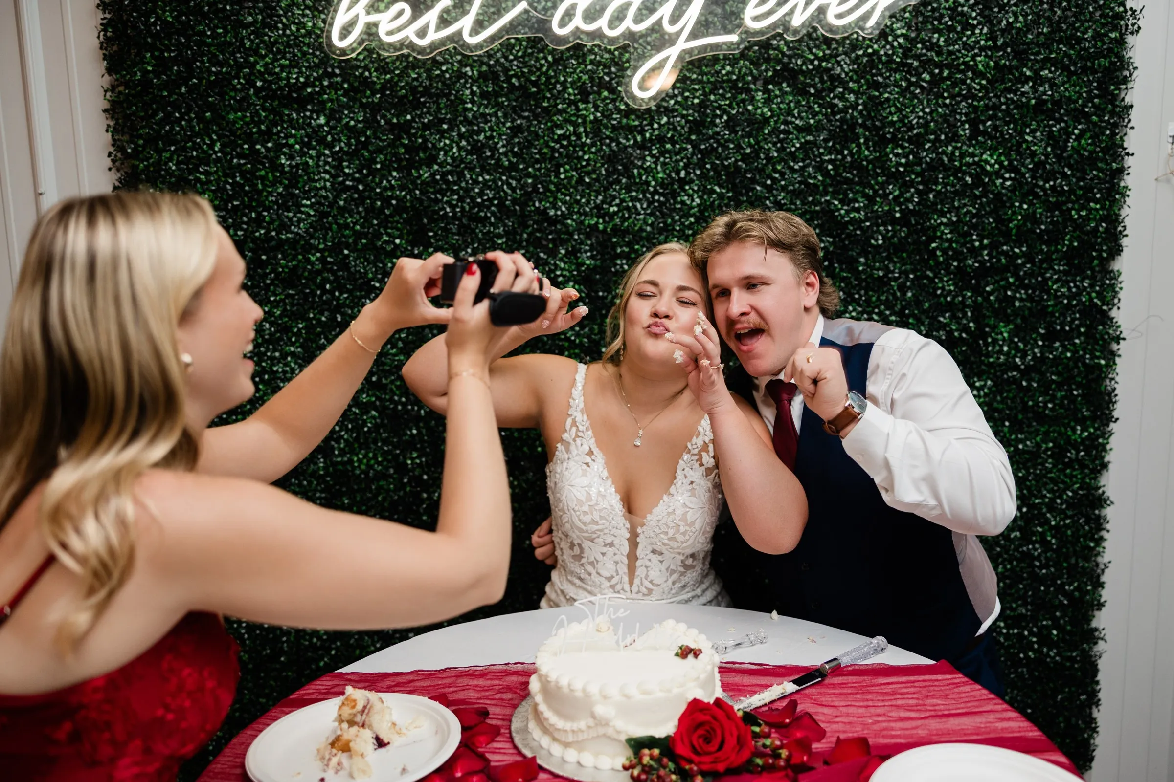 A guest films the couple as they make playful faces with frosting on their fingers beside the cut wedding cake at The Gathering in Crosslake — Tim Larsen Photography, Brainerd Lakes MN