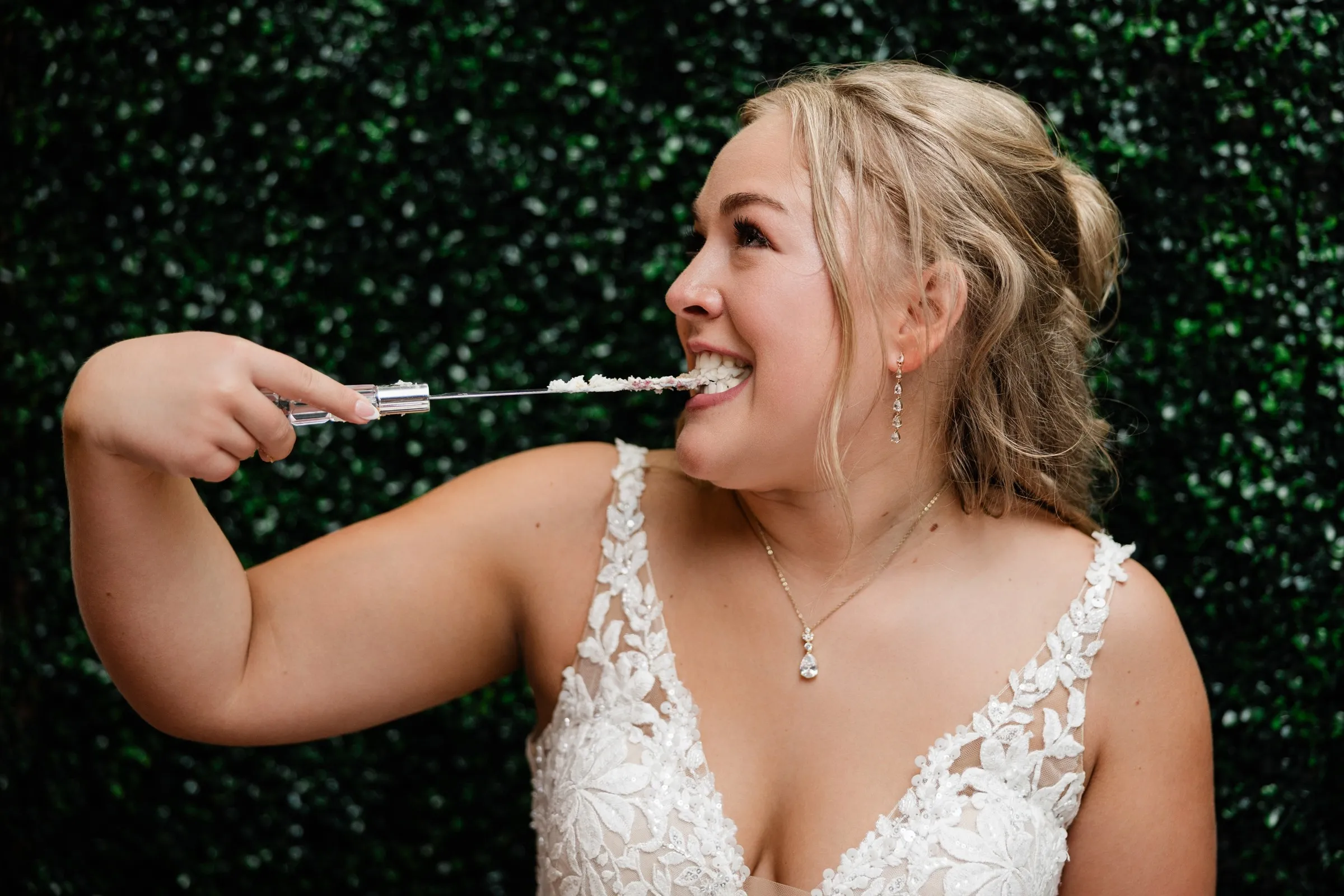 The bride smiles as she tastes frosting from the cake knife in front of the green hedge backdrop at The Gathering in Crosslake — Tim Larsen Photography, Brainerd Lakes MN