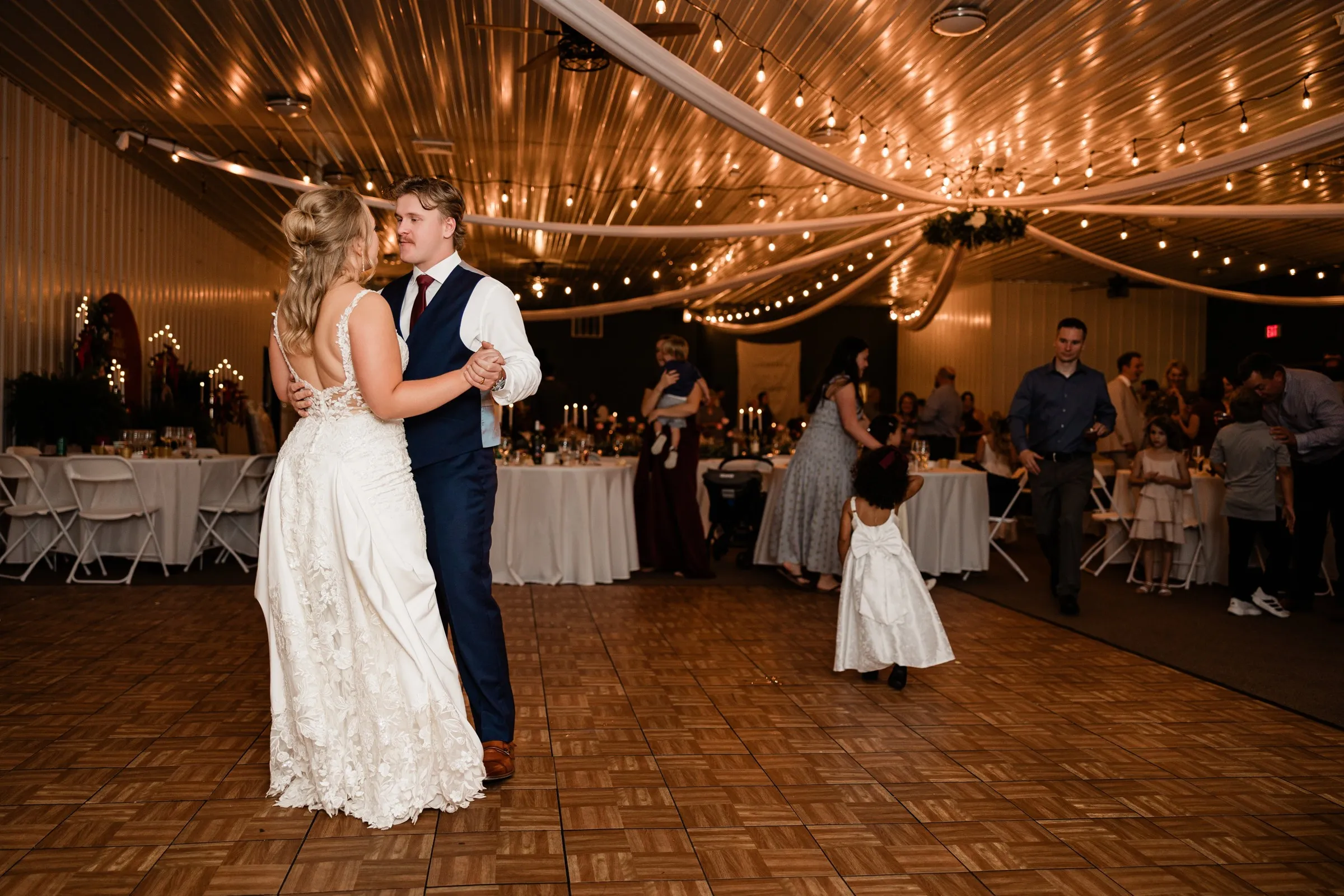 The couple dance on a wooden floor beneath draped fabric and string lights while children and guests gather nearby at The Gathering in Crosslake — Tim Larsen Photography, Brainerd Lakes MN