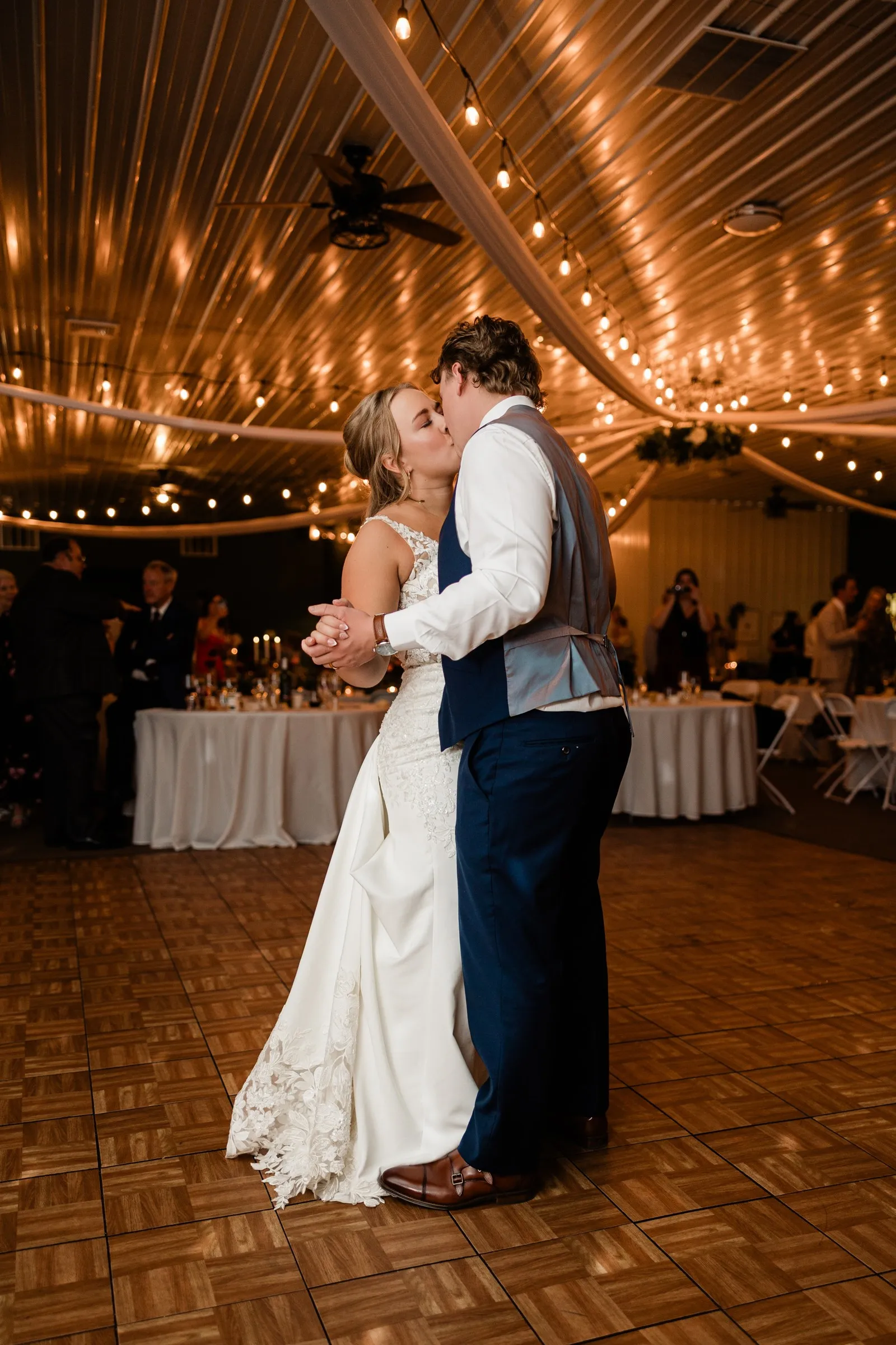 The bride and groom kiss while holding hands on the dance floor beneath warm string lights and white drapery at The Gathering in Crosslake — Tim Larsen Photography, Brainerd Lakes MN