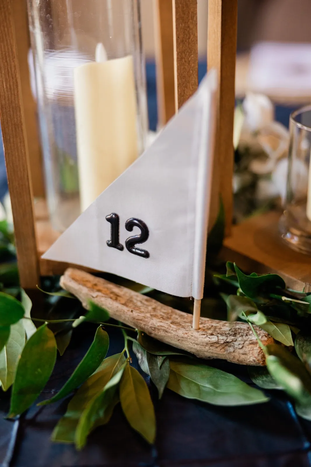 A handmade driftwood sailboat with a white canvas sail marked with the number 12 sits among greenery on a navy table runner beside a wooden lantern holding a pillar candle at Chase on the Lake — Tim Larsen Photography, Brainerd Lakes MN