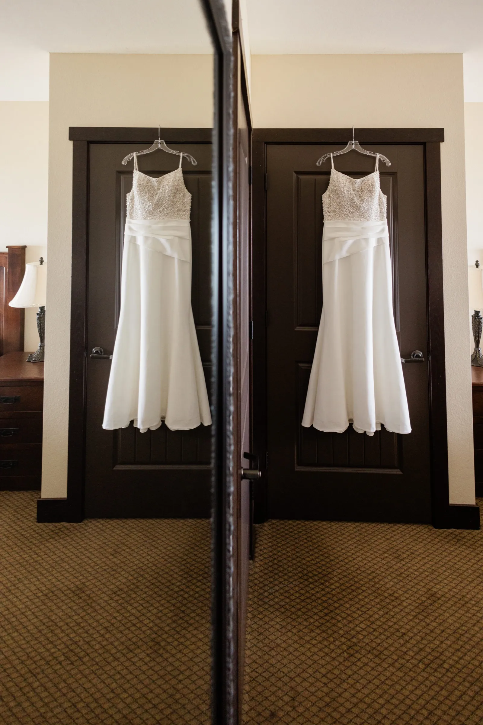 A beaded-bodice wedding gown hangs on a dark hotel door and is doubled by an adjacent mirror in a carpeted guest room — Tim Larsen Photography, Brainerd Lakes MN