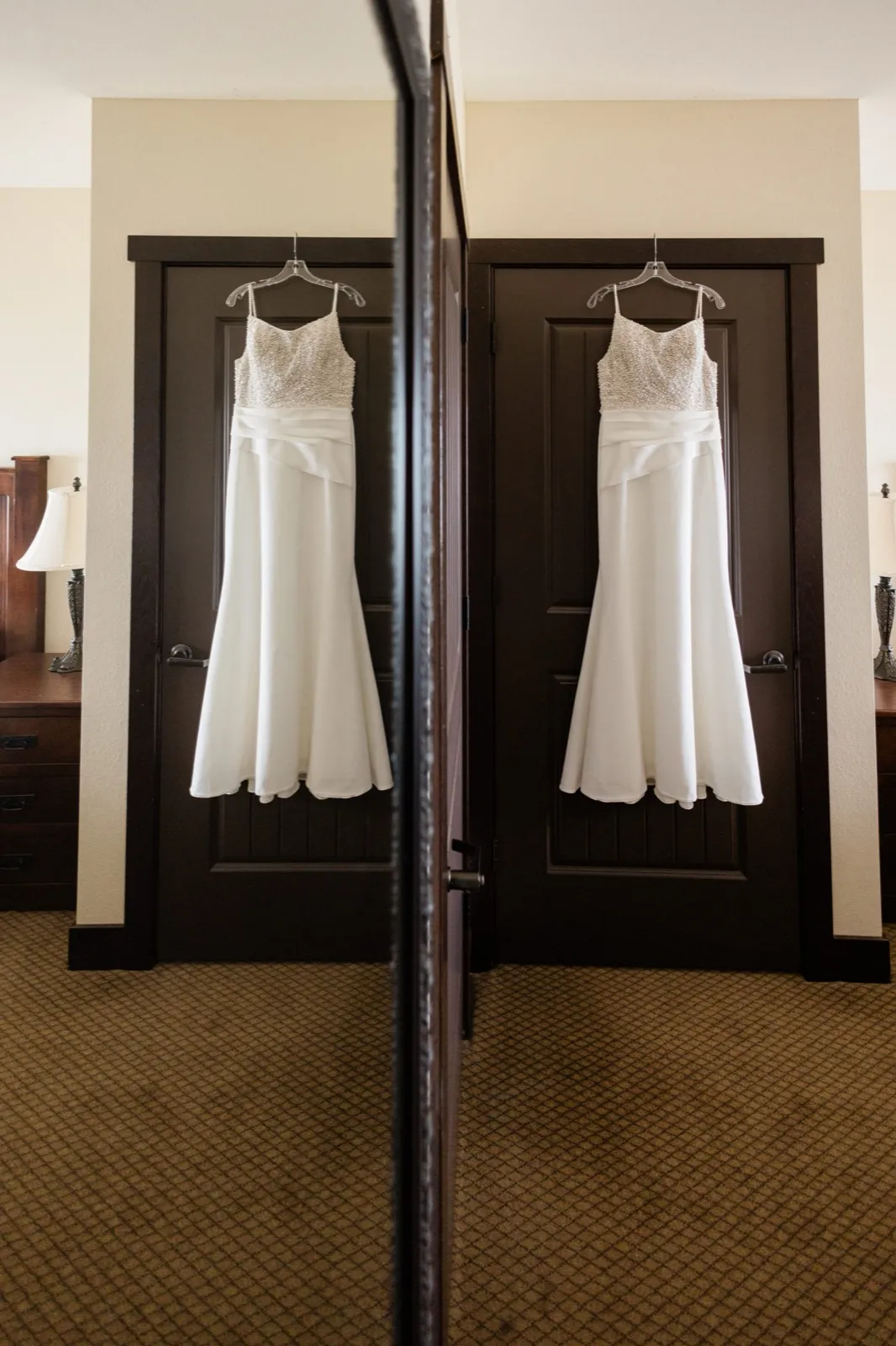 A beaded-bodice wedding gown hangs on a dark hotel door and is doubled by an adjacent mirror in a carpeted guest room — Tim Larsen Photography, Brainerd Lakes MN
