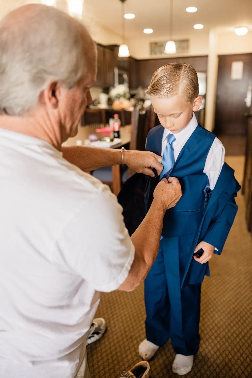 An older man in a white t-shirt adjusts the tie of a young ring bearer in a blue suit inside a resort kitchen and living area — Tim Larsen Photography, Brainerd Lakes MN