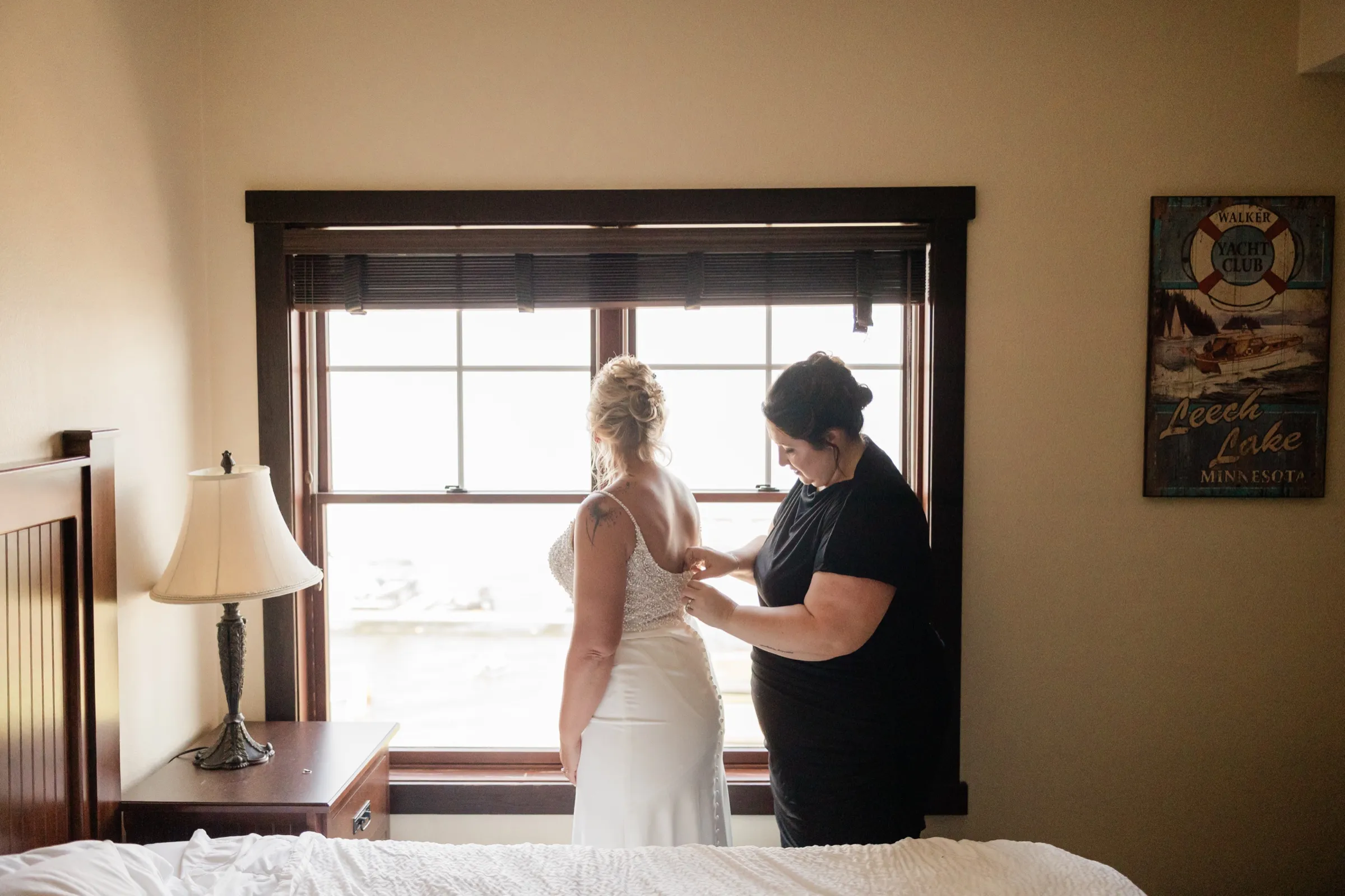 A woman in a black dress fastens the back of the bride's beaded gown as they stand together at a bright window in a hotel room, with a Leech Lake Minnesota sign on the wall to the right — Tim Larsen Photography, Brainerd Lakes MN