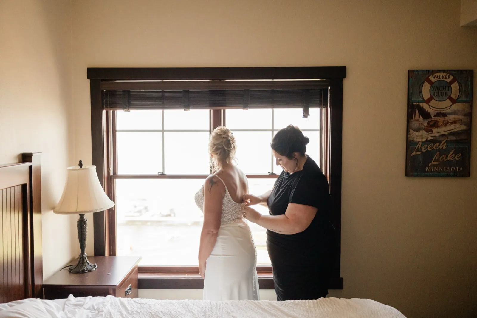 A woman in a black dress fastens the back of the bride's beaded gown as they stand together at a bright window in a hotel room, with a Leech Lake Minnesota sign on the wall to the right — Tim Larsen Photography, Brainerd Lakes MN
