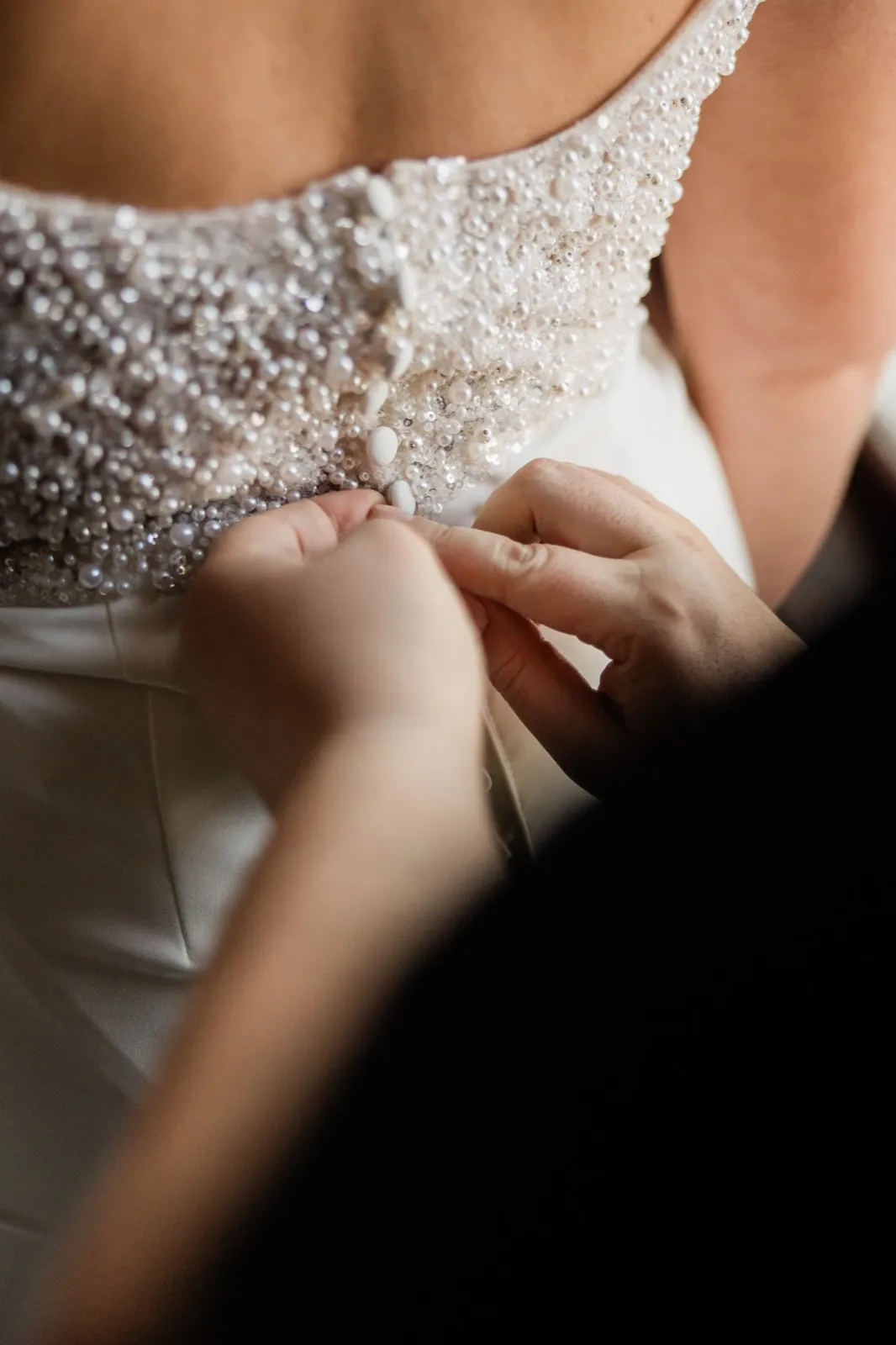Close view of two hands fastening the small buttons on the back of the bride's beaded, pearl-covered gown in soft window light — Tim Larsen Photography, Brainerd Lakes MN