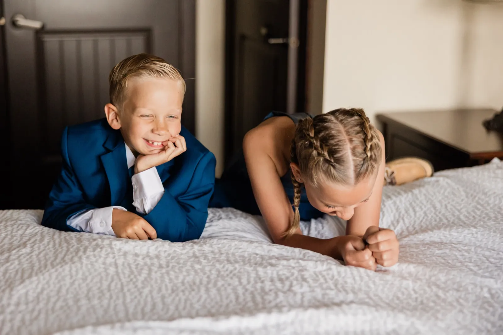 A boy in a blue suit rests his chin on his hand and grins while a girl in a navy dress with braided hair leans down beside him on a white hotel bed — Tim Larsen Photography, Brainerd Lakes MN