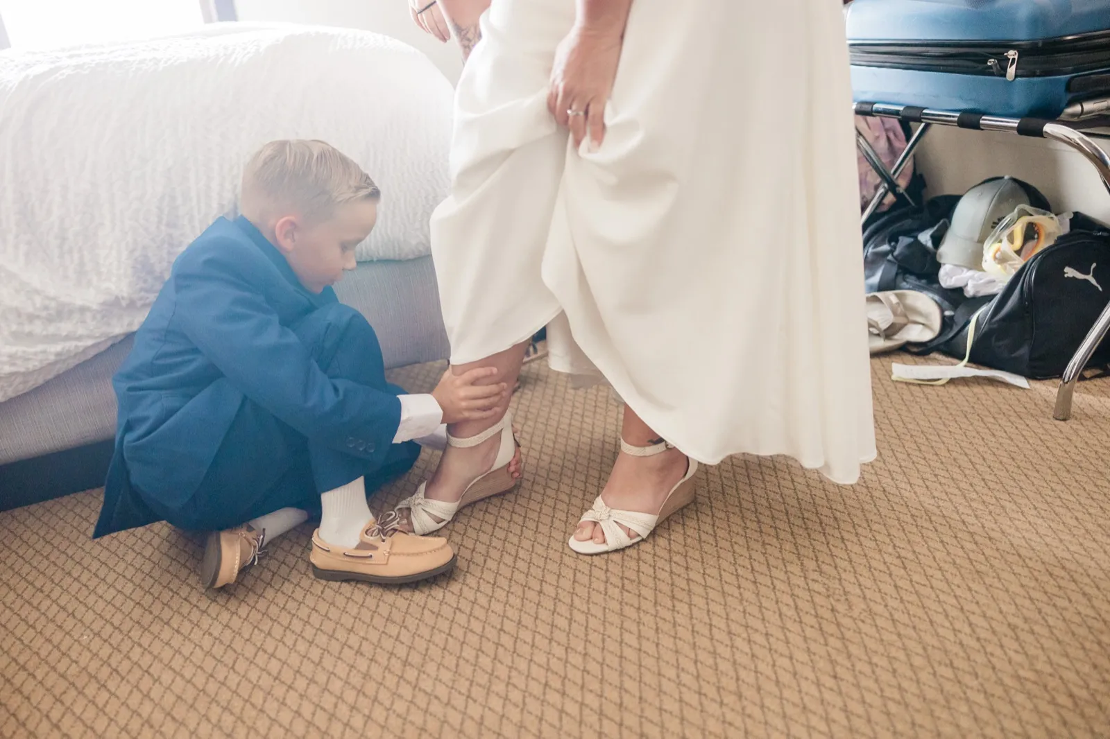A young boy in a blue blazer kneels on the carpet to fasten the strap of the bride's wedge sandal in a hotel room cluttered with luggage — Tim Larsen Photography, Brainerd Lakes MN