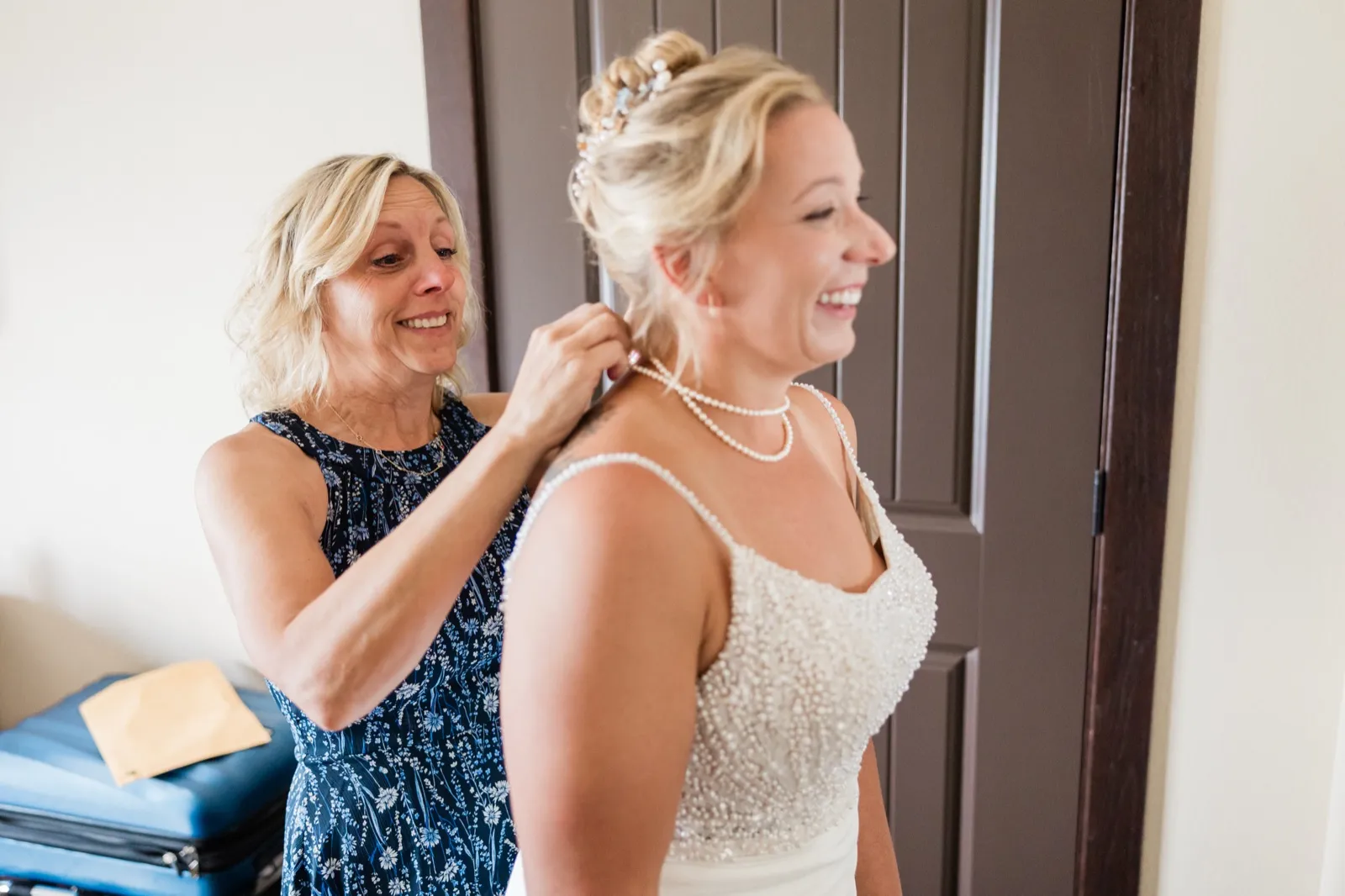 The bride laughs in profile while a family member stands behind her fastening a pearl necklace in a light-filled hotel room — Tim Larsen Photography, Brainerd Lakes MN