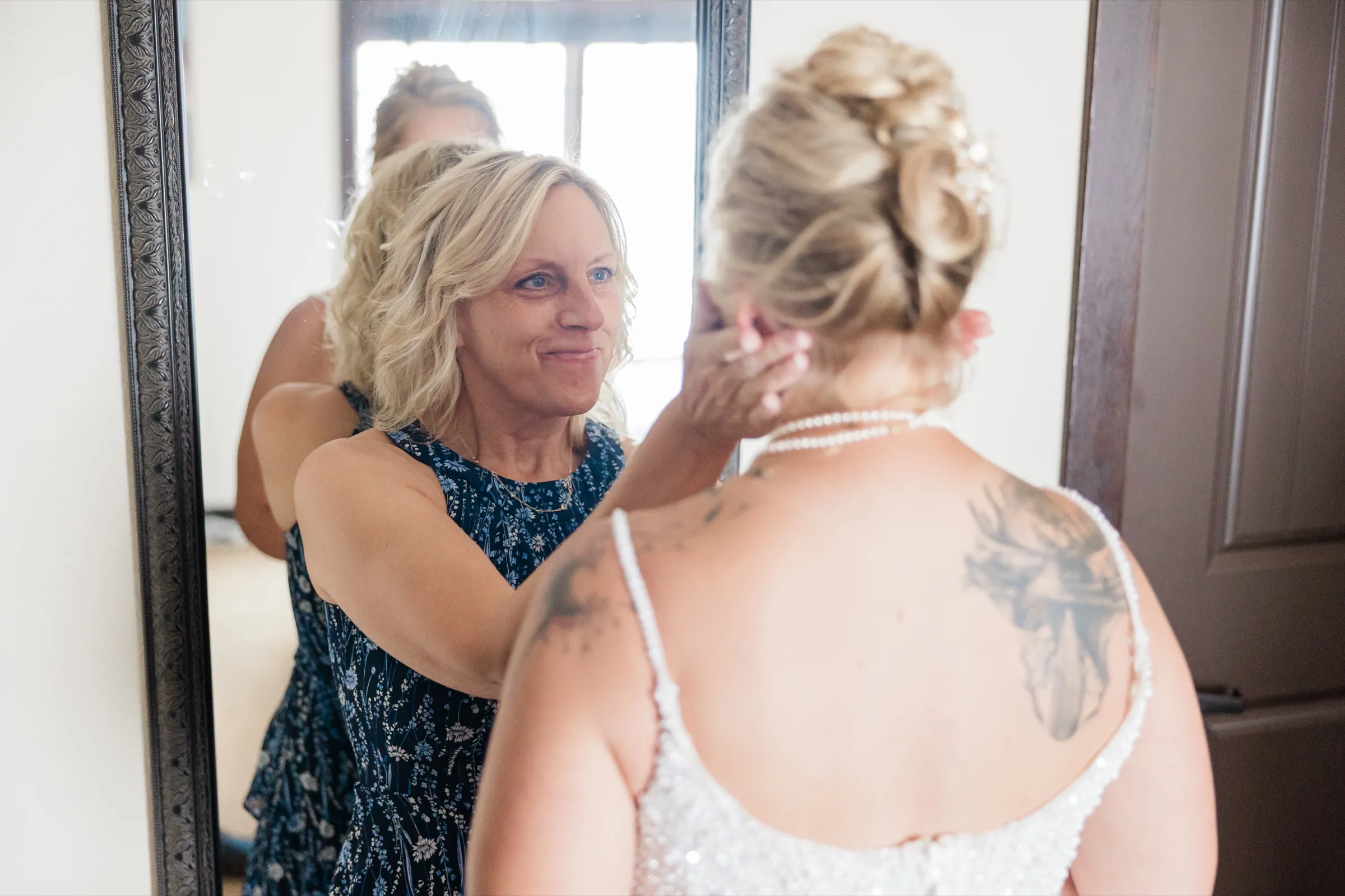 Seen from behind, the bride in a beaded gown has her earring adjusted by a woman in a navy floral dress whose smiling face appears in the mirror ahead of them — Tim Larsen Photography, Brainerd Lakes MN