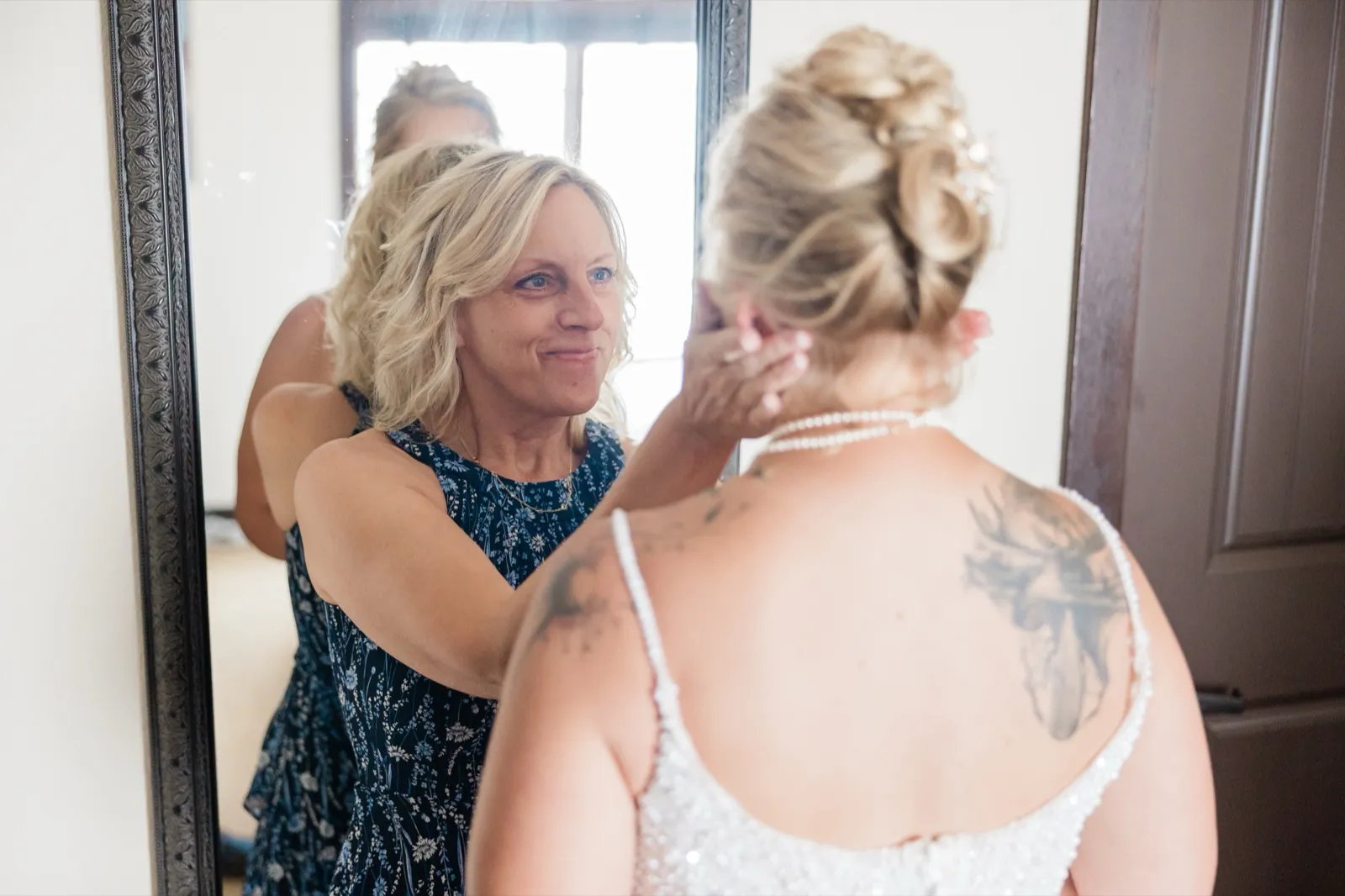 Seen from behind, the bride in a beaded gown has her earring adjusted by a woman in a navy floral dress whose smiling face appears in the mirror ahead of them — Tim Larsen Photography, Brainerd Lakes MN