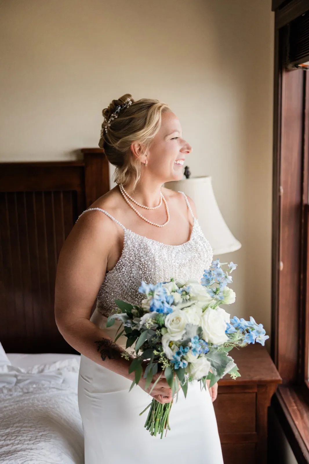 The bride stands in profile beside a window in her hotel room, laughing while holding a white and blue bouquet against her beaded gown — Tim Larsen Photography, Brainerd Lakes MN