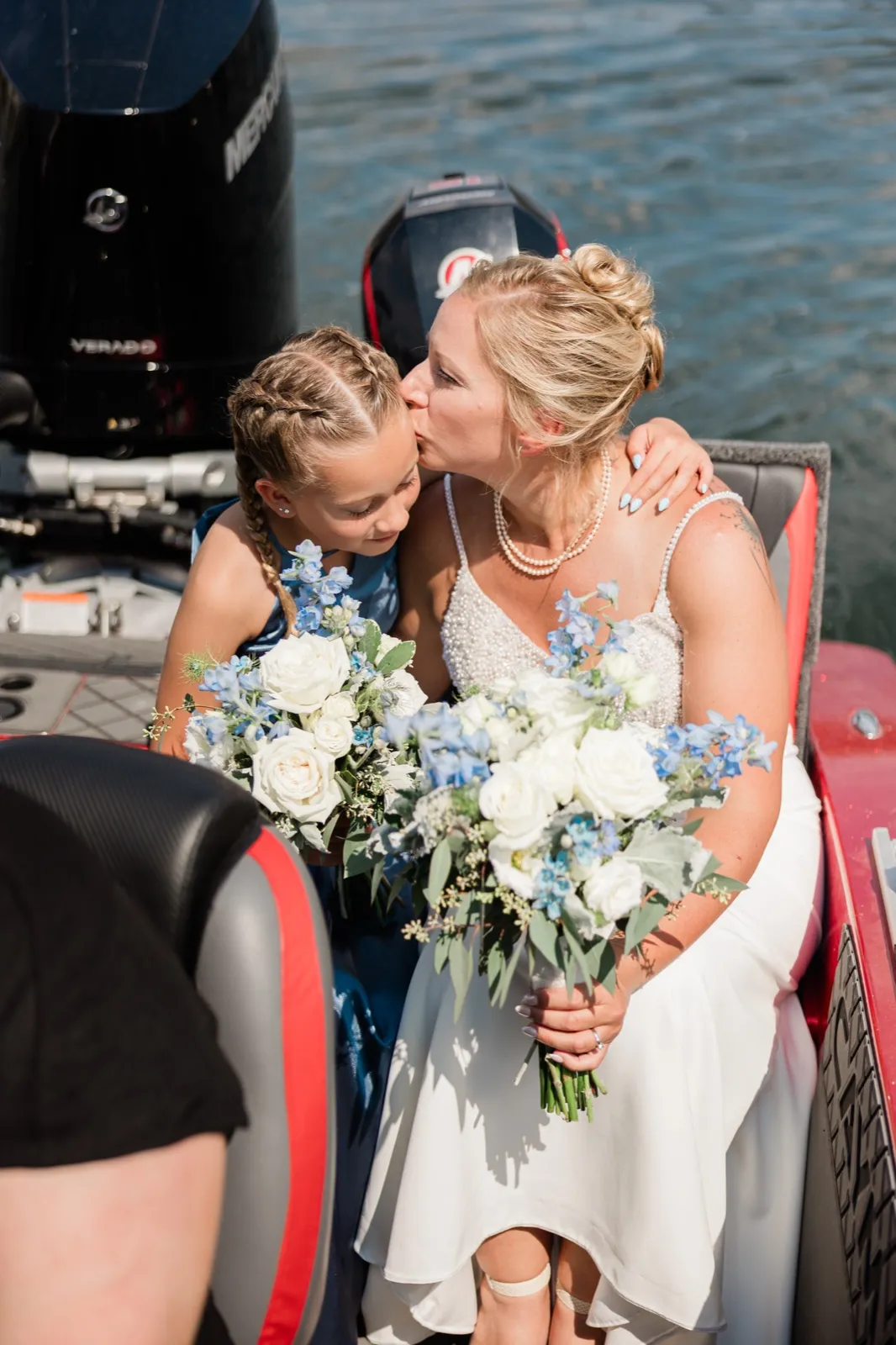 Seated on a boat beside outboard motors, the bride kisses a young girl in a blue dress on the cheek while both hold white and blue bouquets — Tim Larsen Photography, Brainerd Lakes MN