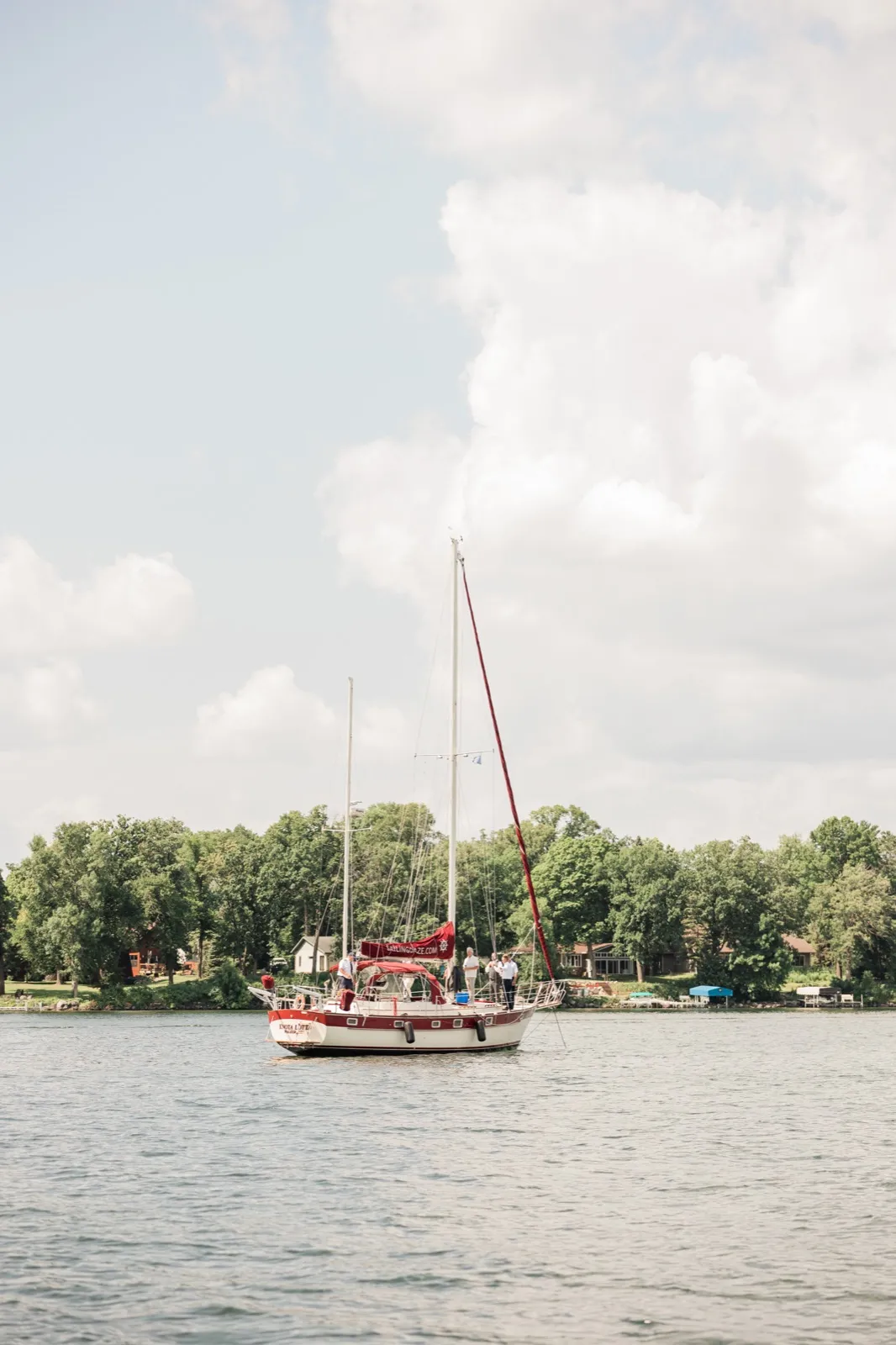 A red and white sailboat carrying a small group of wedding guests sits on Leech Lake beneath a tall summer cloud, with a wooded shoreline behind it — Tim Larsen Photography, Brainerd Lakes MN