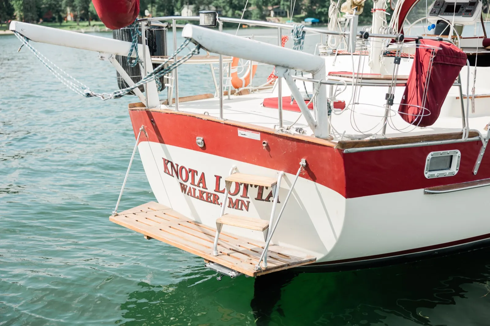 The stern of a red and white sailboat named Knota Lotta out of Walker, Minnesota, floats in green lake water with its wooden swim platform lowered — Tim Larsen Photography, Brainerd Lakes MN