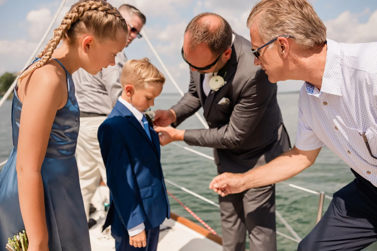 A man in a gray suit pins a white boutonniere to a young boy's navy jacket aboard a sailboat while two other adults and a girl in a blue dress lean in to watch — Tim Larsen Photography, Brainerd Lakes MN
