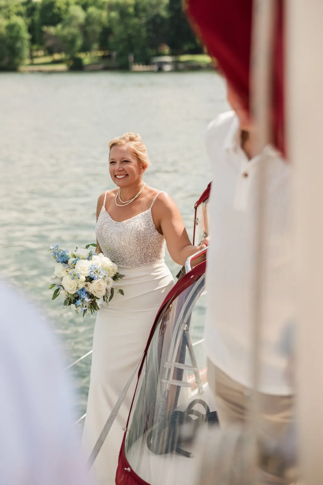 The bride, holding a white and blue bouquet, smiles as she steps aboard a boat on the lake, partly framed by a blurred guest in the foreground — Tim Larsen Photography, Brainerd Lakes MN