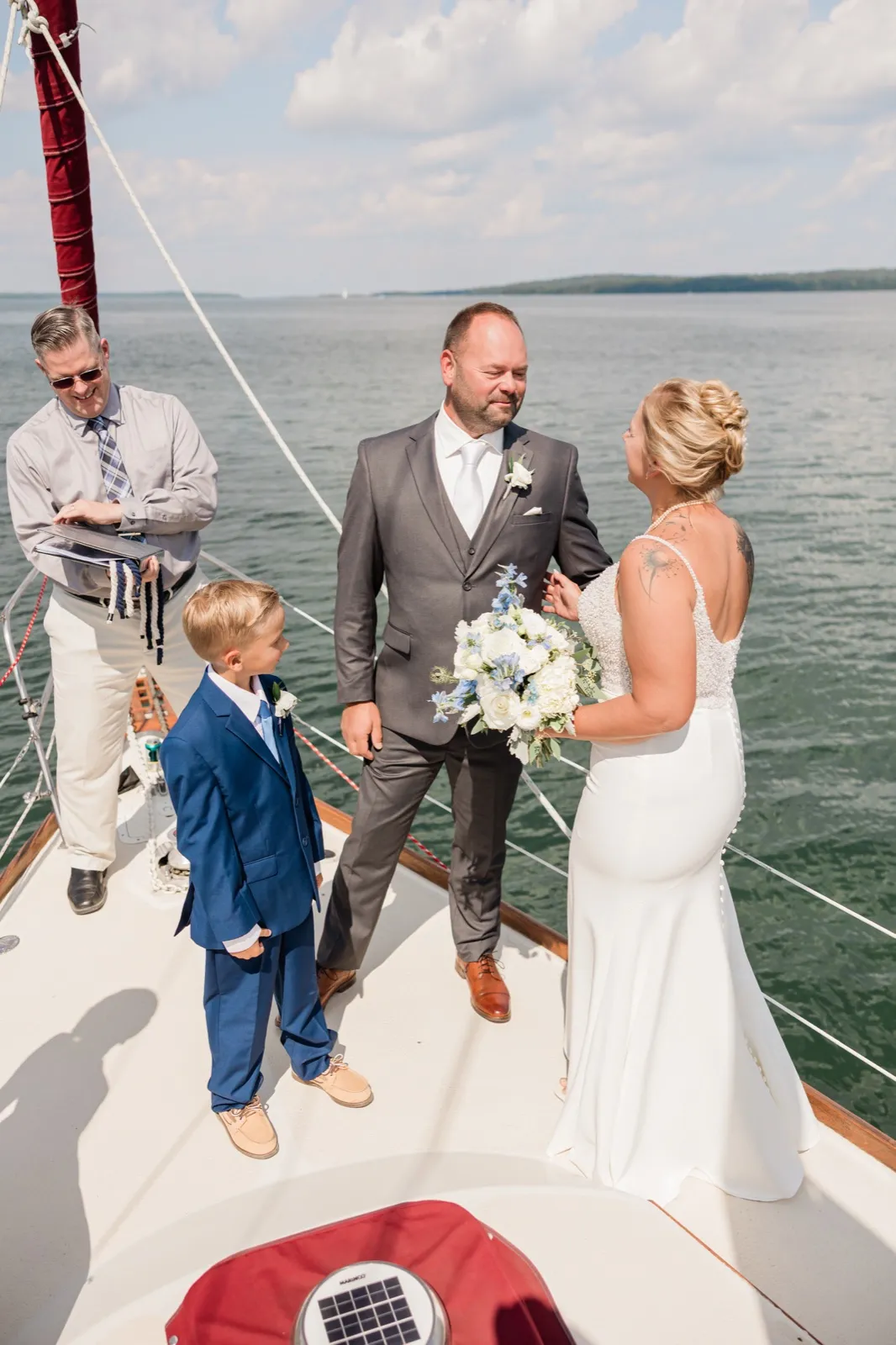 A bride and groom face each other during their ceremony on the bow of a sailboat while an officiant reads from a book and a young boy in a blue suit watches, open lake water behind them — Tim Larsen Photography, Brainerd Lakes MN
