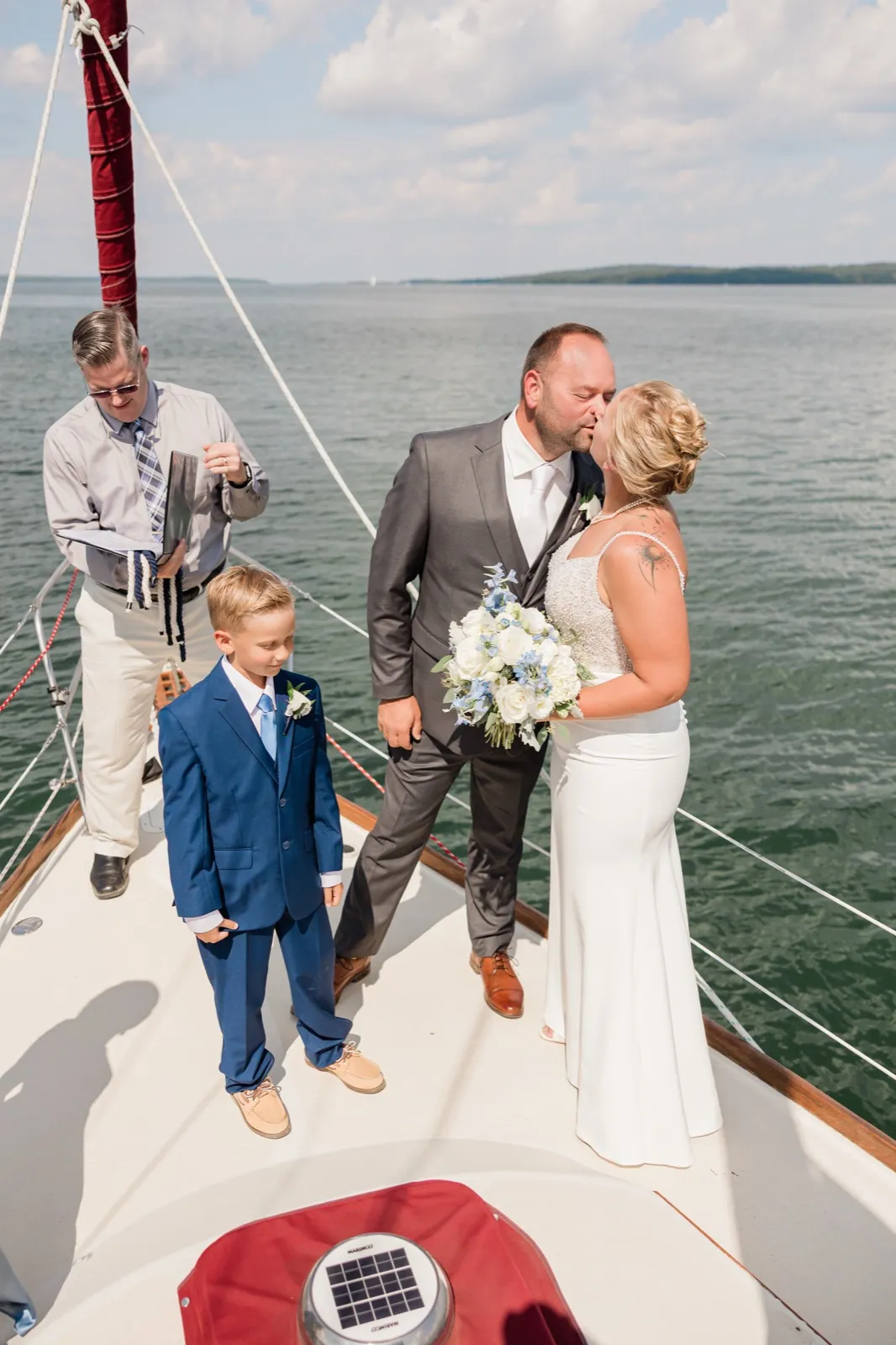 The bride and groom kiss on the bow of a sailboat while a young boy in a blue suit stands beside them and the officiant closes his book, open lake water behind them — Tim Larsen Photography, Brainerd Lakes MN