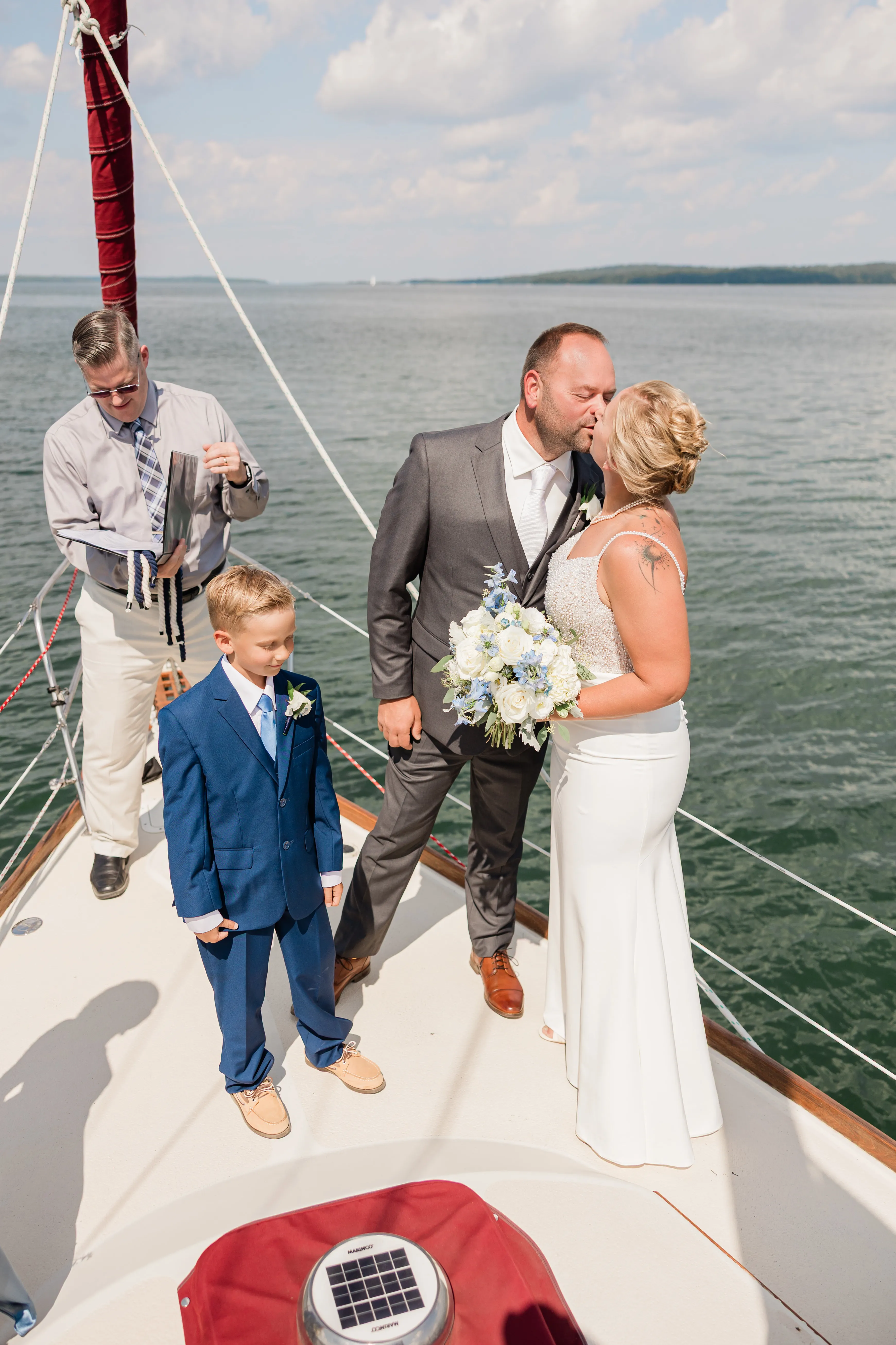 The bride and groom kiss on the bow of a sailboat while a young boy in a blue suit stands beside them and the officiant closes his book, open lake water behind them — Tim Larsen Photography, Brainerd Lakes MN