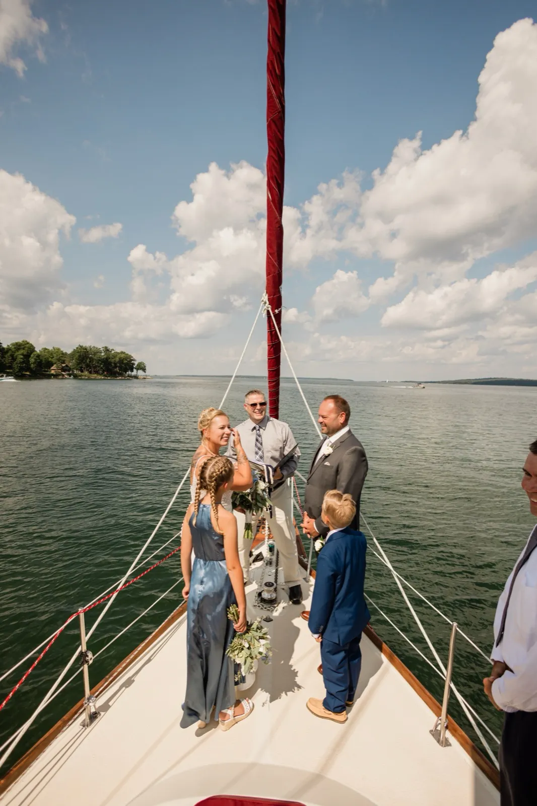 The bride and groom stand facing each other on the bow of a sailboat as an officiant reads from a book, with two children in the wedding party looking on and open lake water and clouds behind them — Tim Larsen Photography, Brainerd Lakes MN