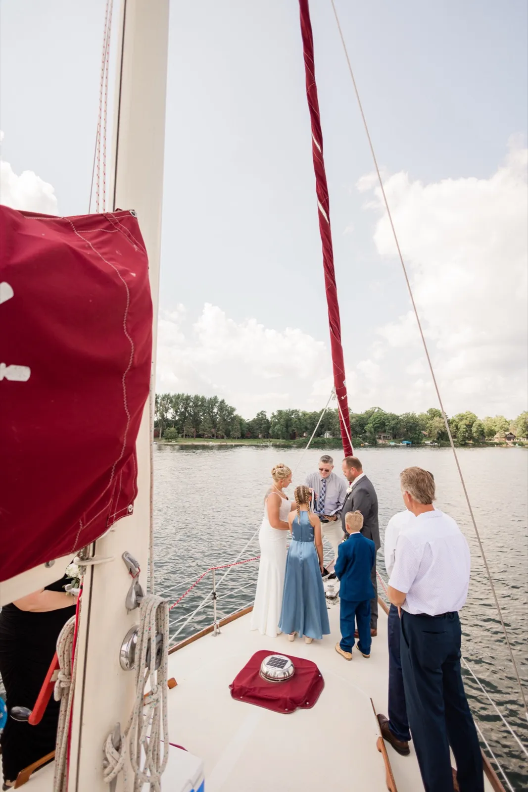 A small wedding party stands on the bow of a sailboat as the officiant reads, with a crimson sail cover and rigging in the foreground and a tree-lined lakeshore behind them — Tim Larsen Photography, Brainerd Lakes MN