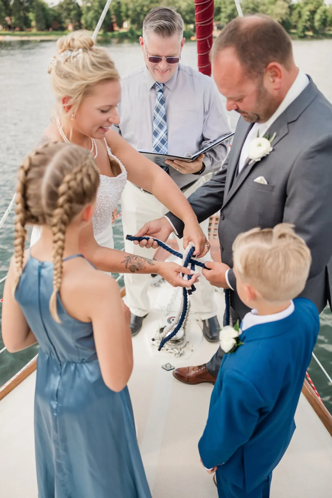 The bride and groom tie a navy and white sailor's knot during their ceremony aboard a sailboat while the officiant reads and two children watch from the foreground — Tim Larsen Photography, Brainerd Lakes MN