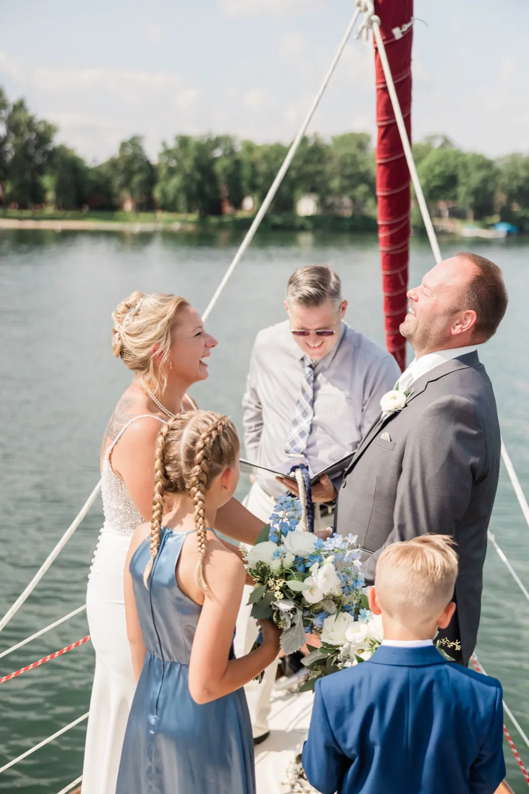 The bride and groom throw their heads back laughing during their ceremony aboard a sailboat, with the officiant reading behind them and two children holding a blue-and-white bouquet in the foreground — Tim Larsen Photography, Brainerd Lakes MN