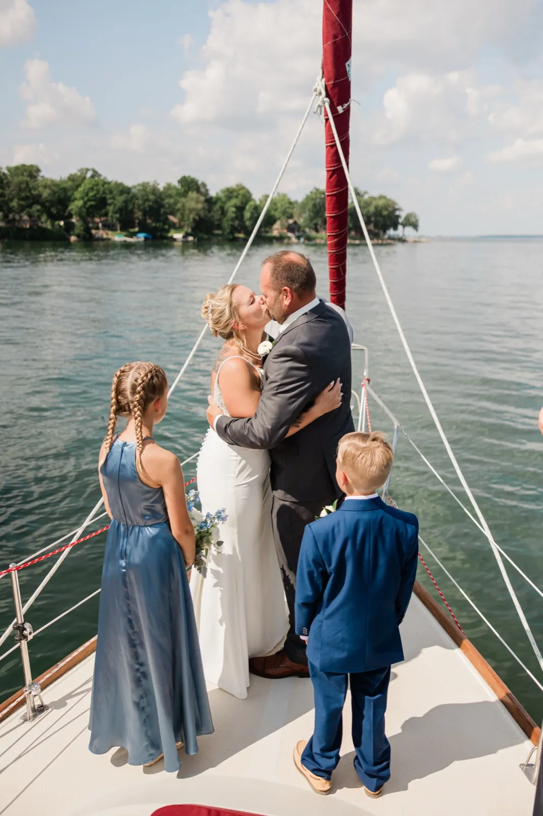 The bride and groom kiss on the bow of a sailboat on open water while a young girl in a blue dress and a boy in a navy suit watch from either side — Tim Larsen Photography, Brainerd Lakes MN