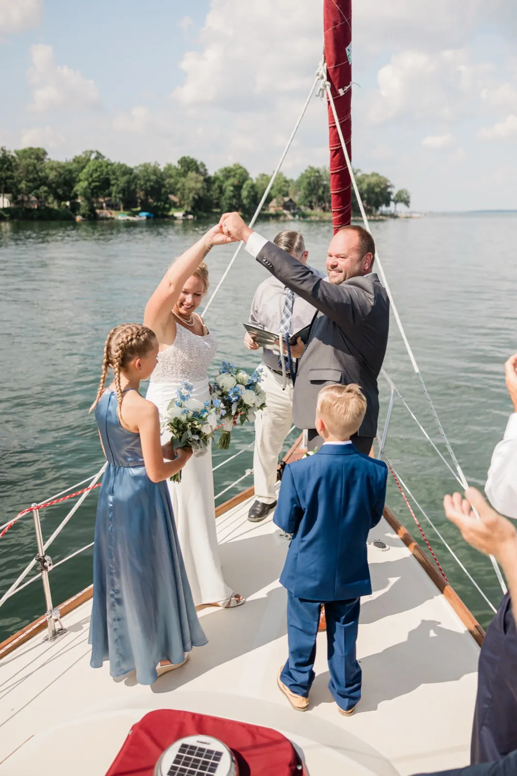 The bride and groom raise their joined hands in celebration on the bow of a sailboat while a young flower girl and ring bearer watch and the officiant stands behind them on the water — Tim Larsen Photography, Brainerd Lakes MN
