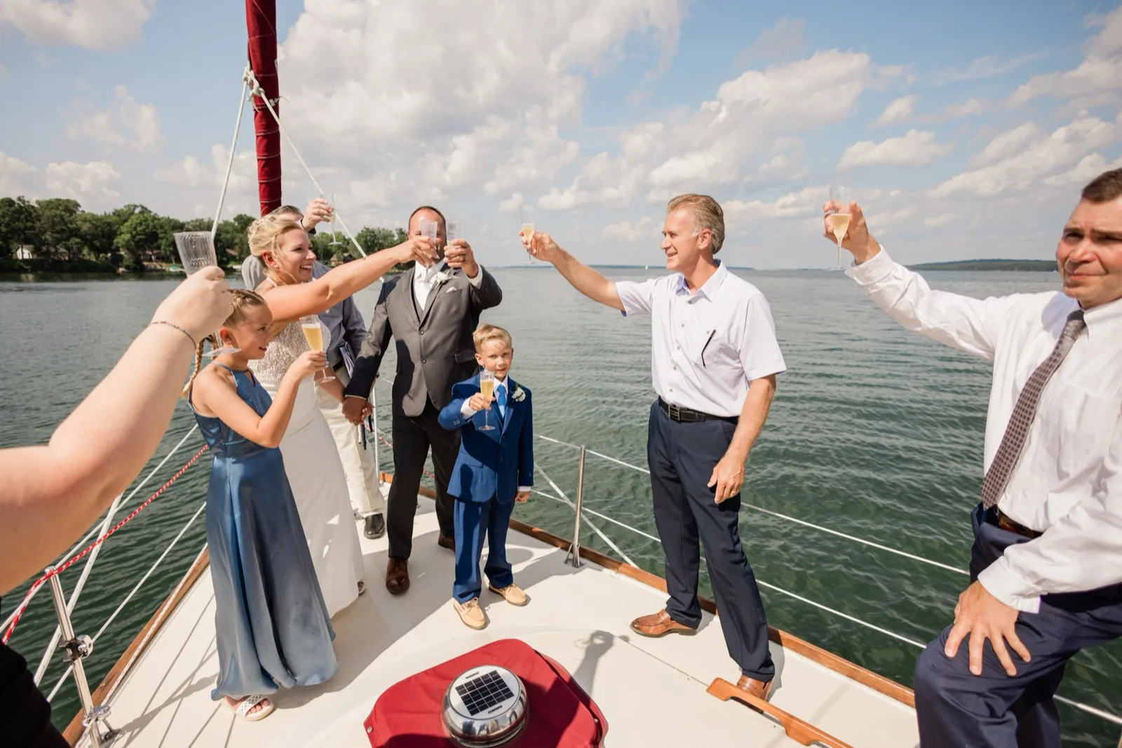 Guests and family raise champagne glasses toward the bride and groom on the deck of a sailboat, with open lake water and clouds behind them — Tim Larsen Photography, Brainerd Lakes MN