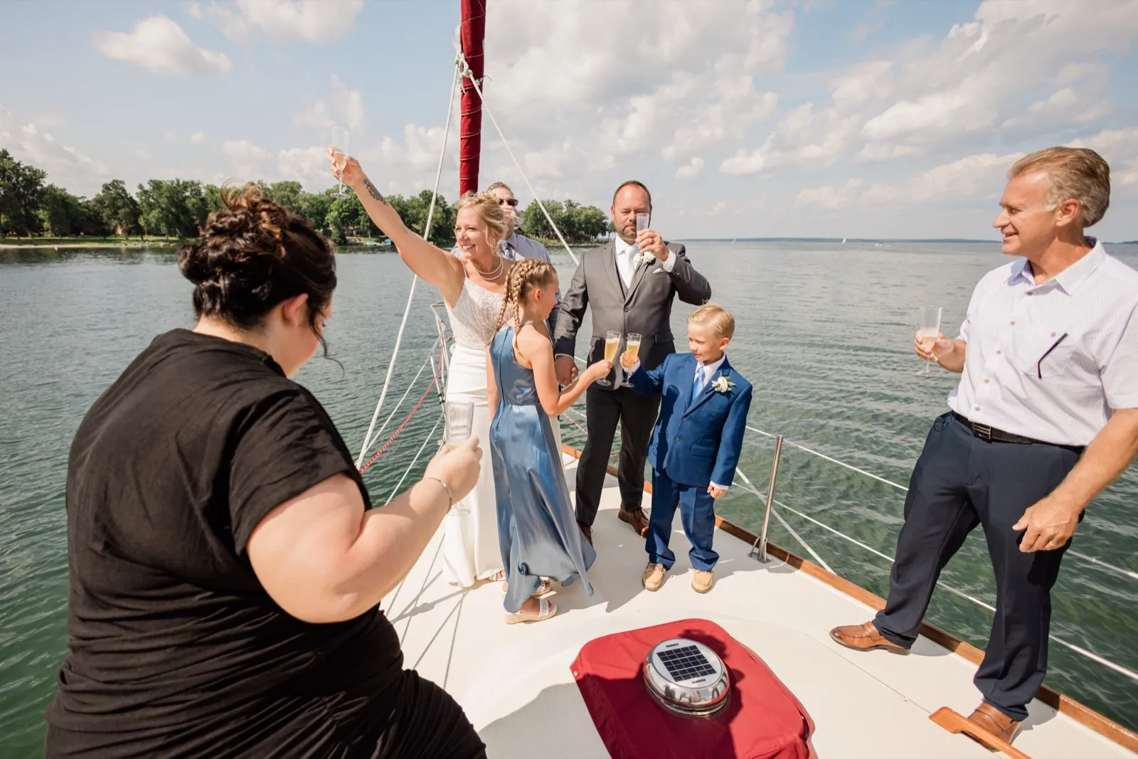 The bride raises a champagne glass overhead while the groom and two children toast on the bow of a sailboat surrounded by open lake water — Tim Larsen Photography, Brainerd Lakes MN