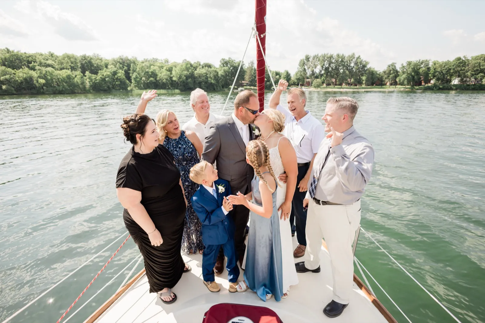 The bride and groom kiss on the bow of a sailboat while nine family members and guests around them cheer with raised arms, the green shoreline of Leech Lake behind them — Tim Larsen Photography, Brainerd Lakes MN