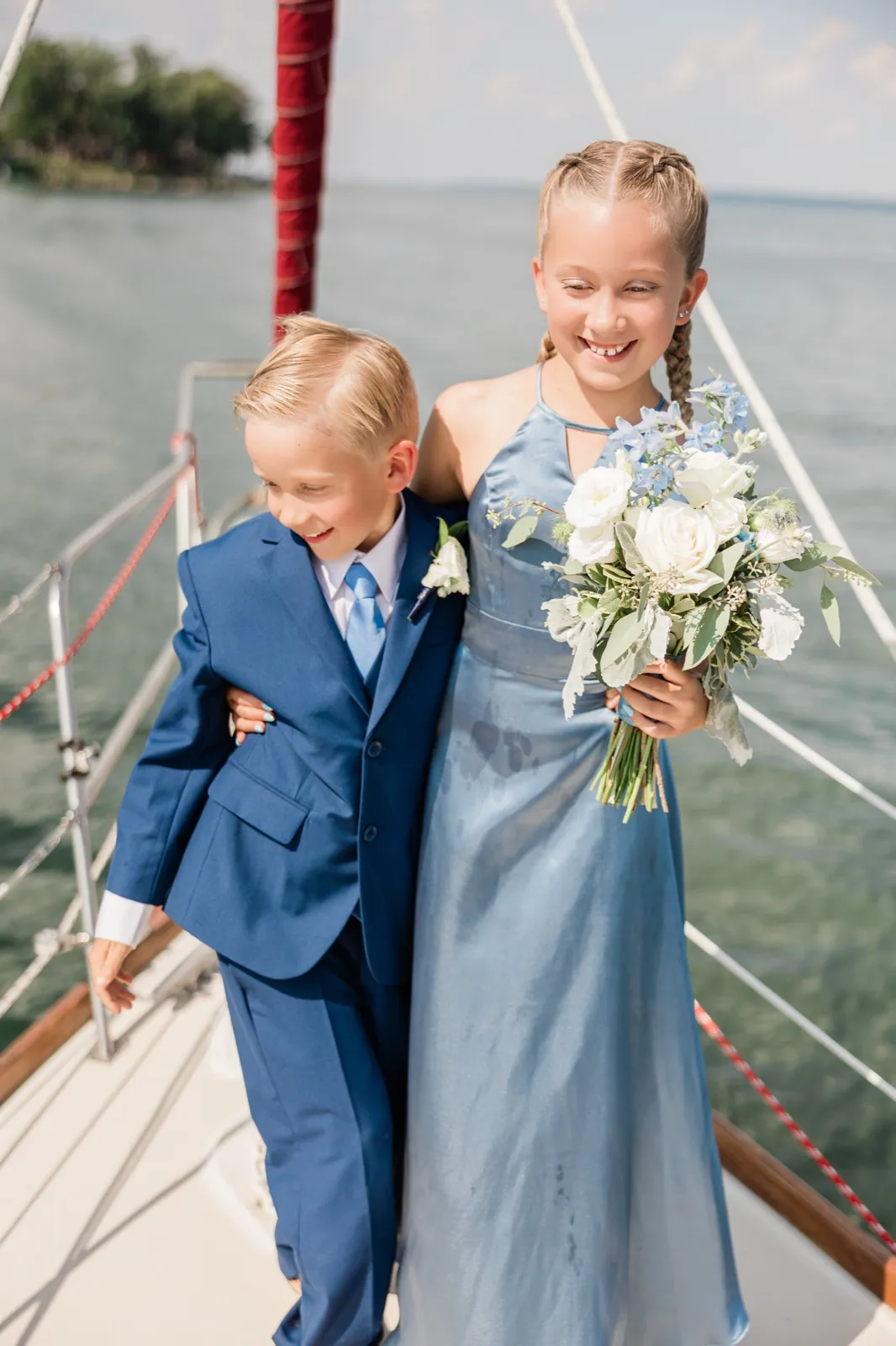 A young flower girl in a dusty blue dress holds a white and blue bouquet while walking arm in arm with a young boy in a navy suit along the deck of a sailboat on a lake — Tim Larsen Photography, Brainerd Lakes MN