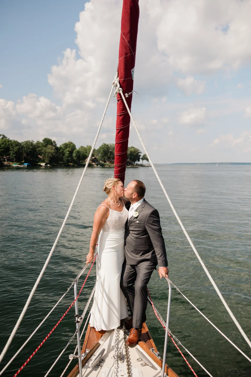 The bride and groom kiss on the bow of a sailboat, framed by white rigging lines and a furled red sail, with Leech Lake and a wooded shoreline behind them — Tim Larsen Photography, Brainerd Lakes MN