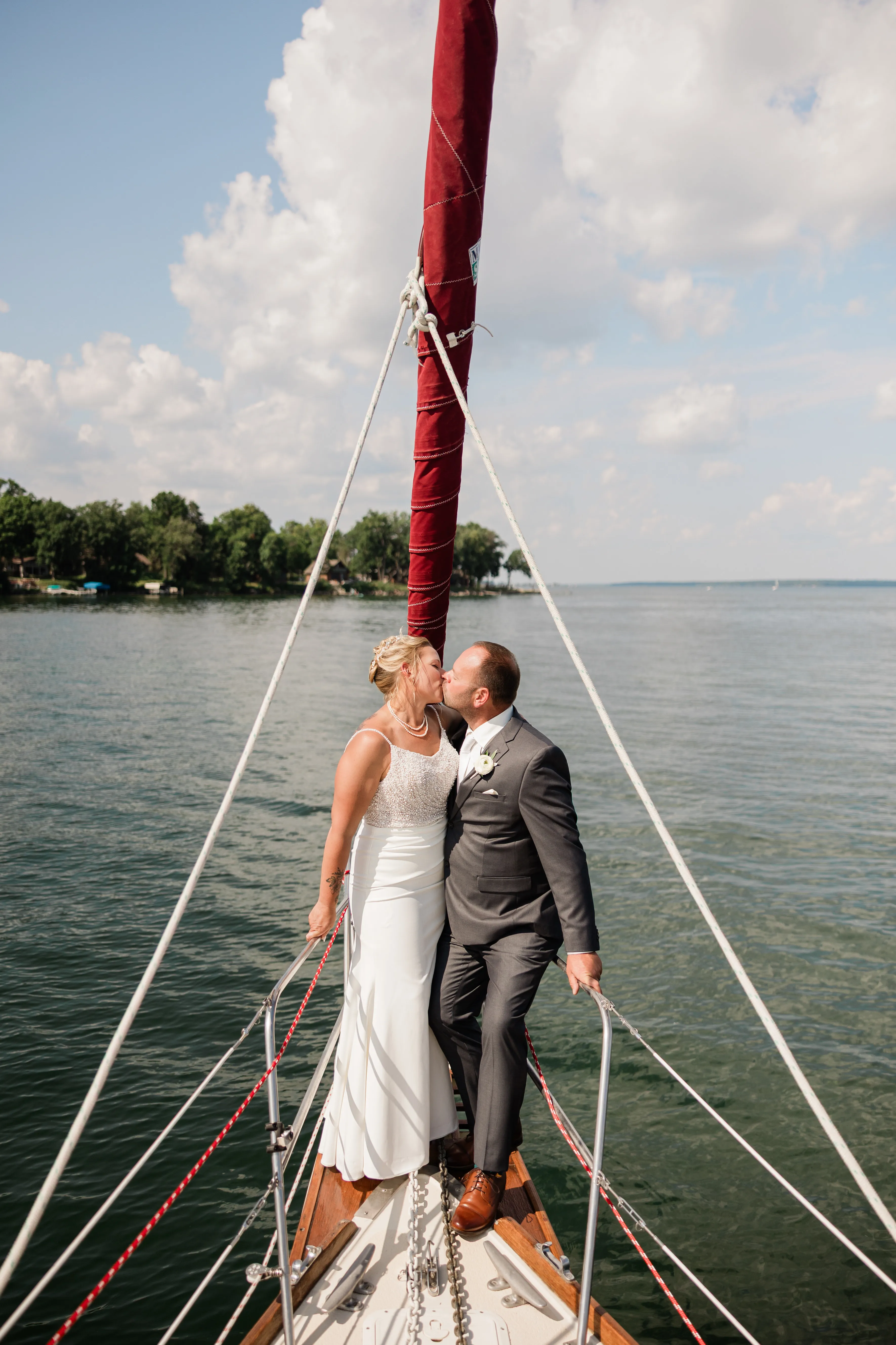 The bride and groom kiss on the bow of a sailboat, framed by white rigging lines and a furled red sail, with Leech Lake and a wooded shoreline behind them — Tim Larsen Photography, Brainerd Lakes MN