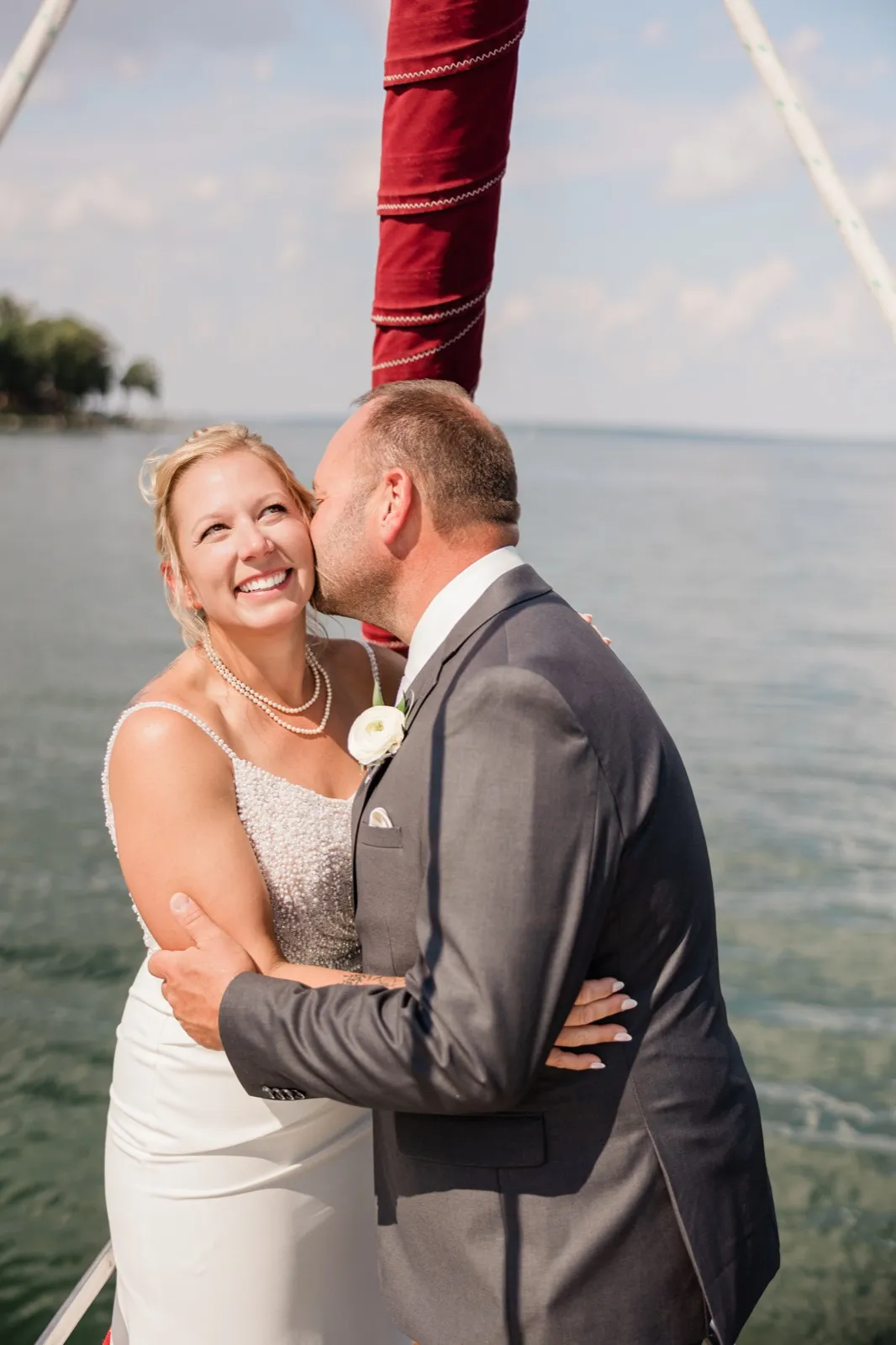 The groom kisses the bride's cheek as she smiles and looks upward, the two embracing on a sailboat deck with open lake water behind them — Tim Larsen Photography, Brainerd Lakes MN