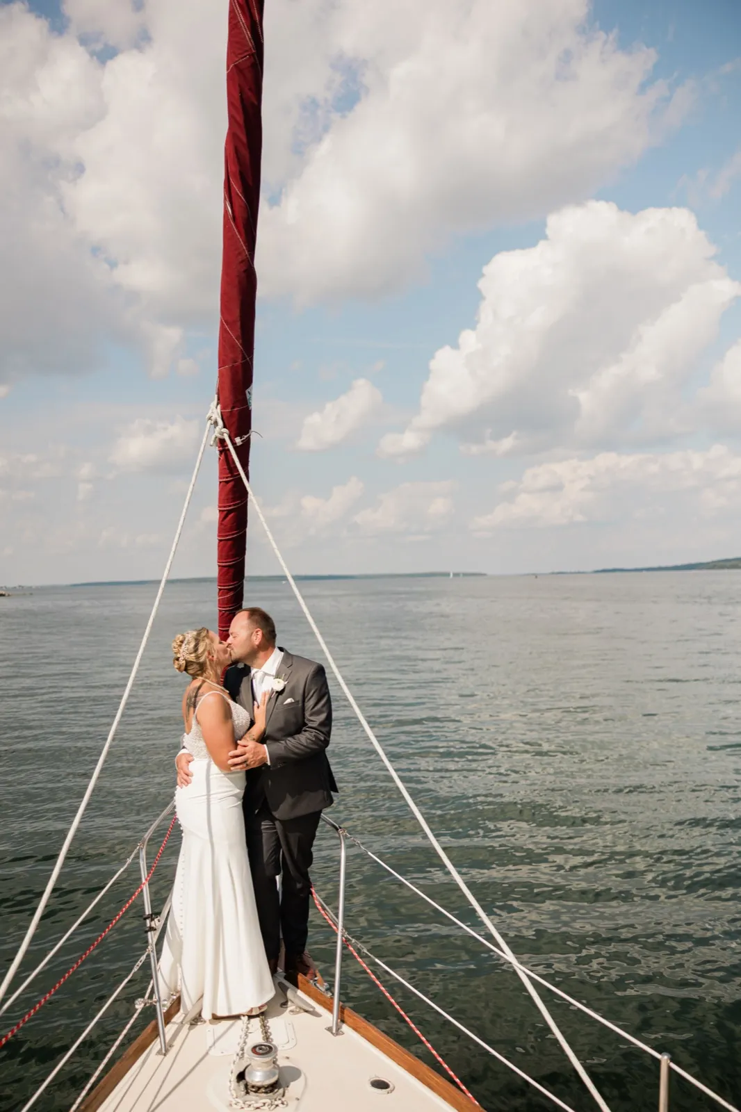 The bride and groom kiss at the bow of a sailboat on open water, framed by white rigging lines and a tall furled sail against a cloud-filled sky — Tim Larsen Photography, Brainerd Lakes MN