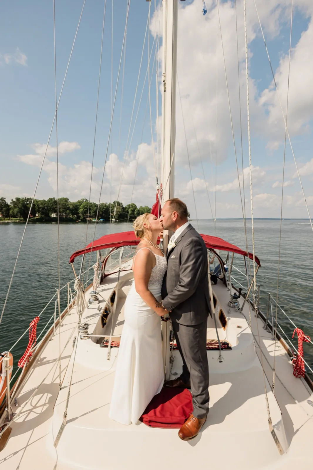 The bride and groom kiss on the white foredeck of a sailboat, surrounded by rigging lines with Leech Lake and a wooded shoreline behind them — Tim Larsen Photography, Brainerd Lakes MN