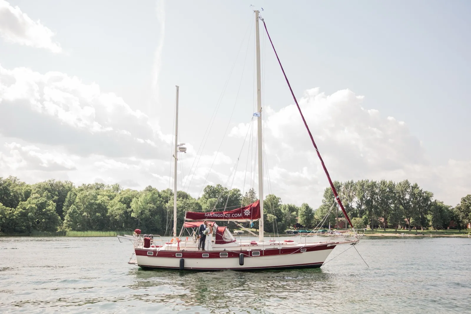The bride and groom kiss on the deck of a red-and-white sailboat anchored on Leech Lake, with a wooded shoreline and tall summer clouds behind them — Tim Larsen Photography, Brainerd Lakes MN
