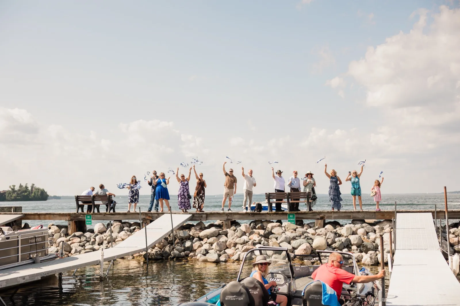 About twenty wedding guests stand in a line along a long lake dock waving blue and white ribbon wands toward the water, with boats and a rocky shoreline in the foreground — Tim Larsen Photography, Brainerd Lakes MN