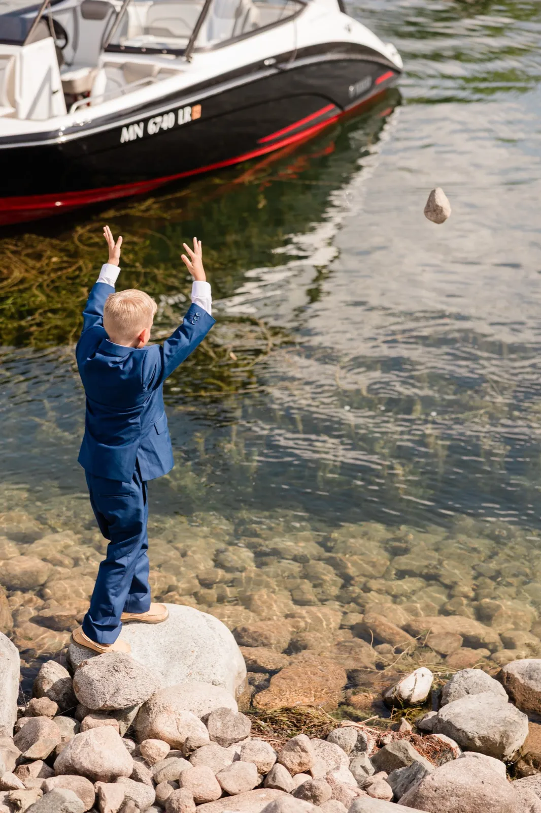 A young boy in a navy suit stands on a rock at the edge of a clear lake, arms raised after tossing a stone that hangs mid-air above the water, with a moored boat behind him — Tim Larsen Photography, Brainerd Lakes MN