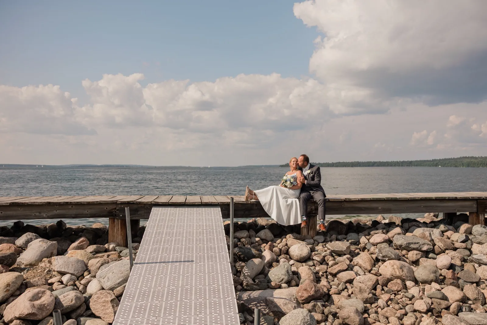The bride and groom sit together at the edge of a wooden dock above a rocky shoreline, her legs swung up beside her as he leans in toward her cheek, with Leech Lake and a tall cloud bank behind them — Tim Larsen Photography, Brainerd Lakes MN