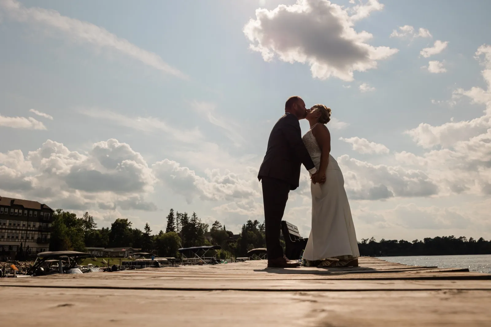 The bride and groom kiss while standing at the end of a wooden dock on Leech Lake, backlit by afternoon sun with a single cloud overhead — Tim Larsen Photography, Brainerd Lakes MN