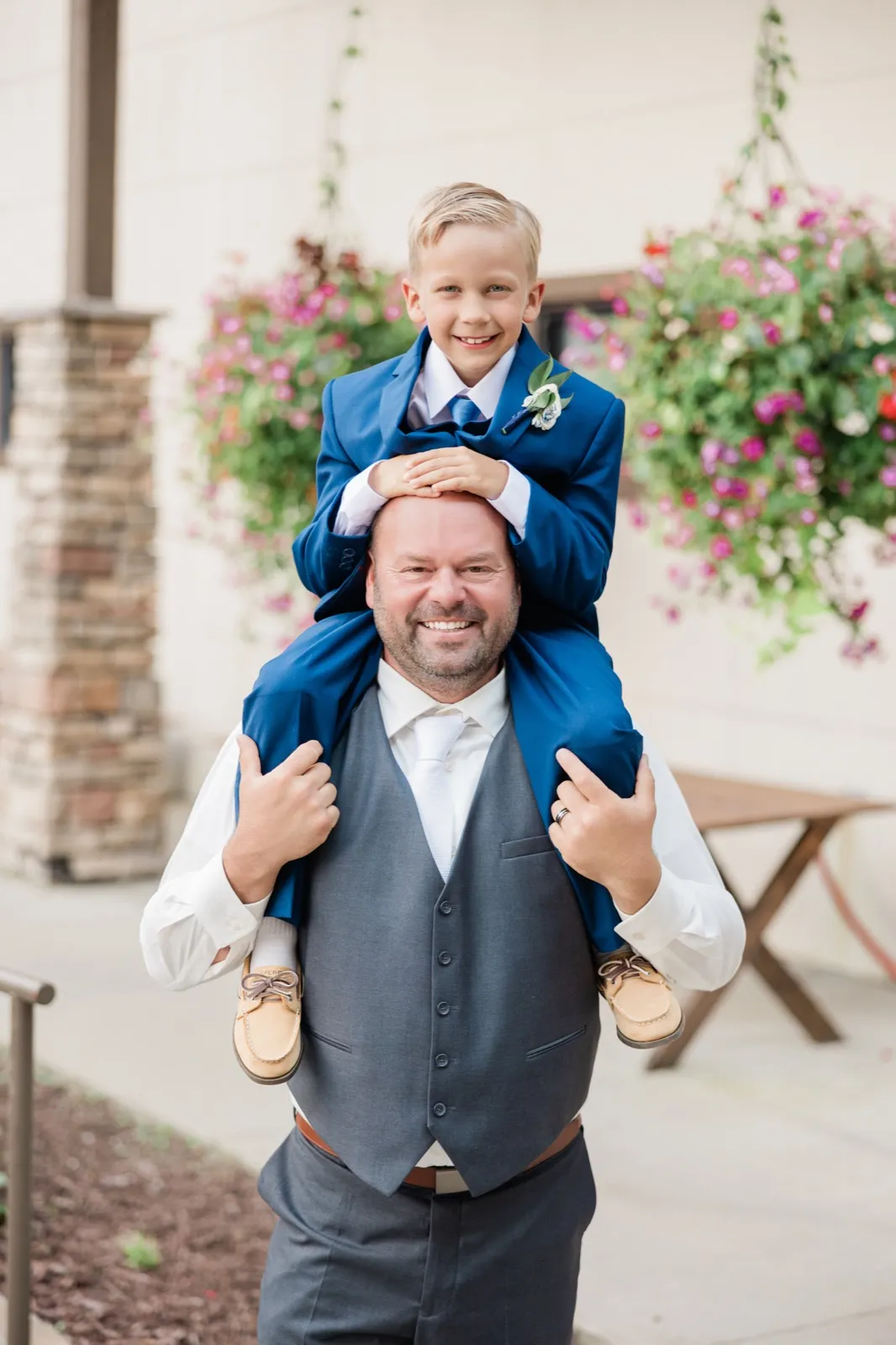 A young boy in a blue suit sits on a man's shoulders, both smiling at the camera on a resort patio with hanging flower baskets behind them — Tim Larsen Photography, Brainerd Lakes MN