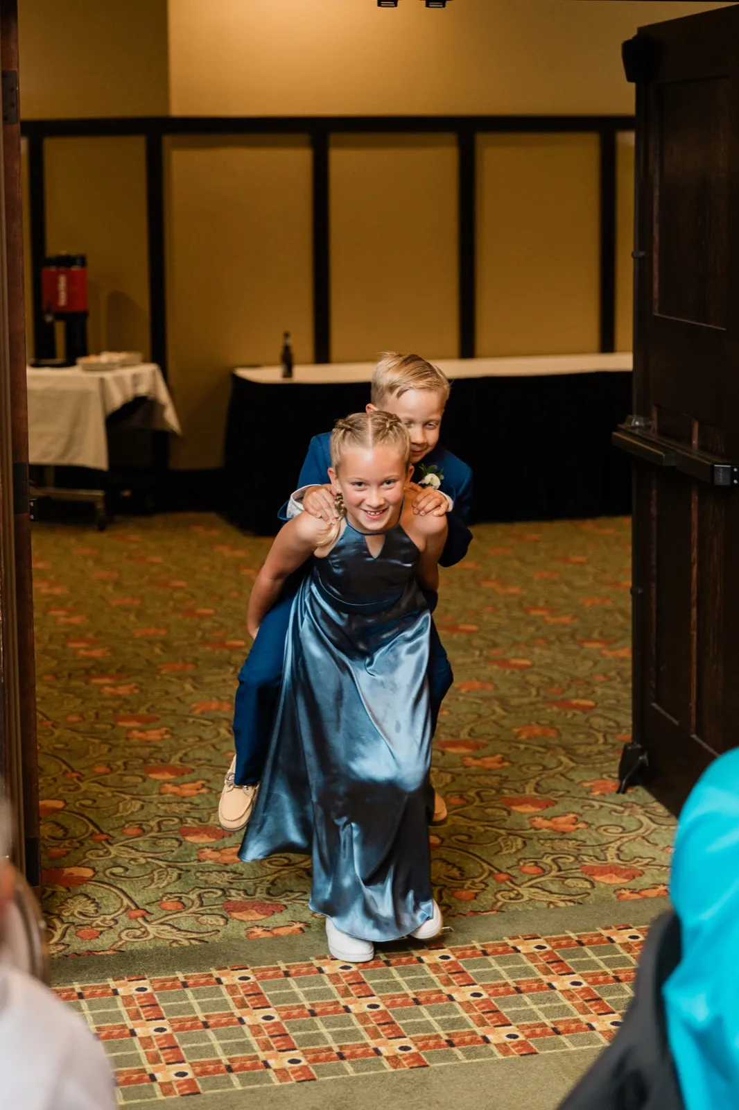 A young girl in a blue satin dress carries a boy in a navy suit on her back as they enter a reception hall through an open doorway — Tim Larsen Photography, Brainerd Lakes MN