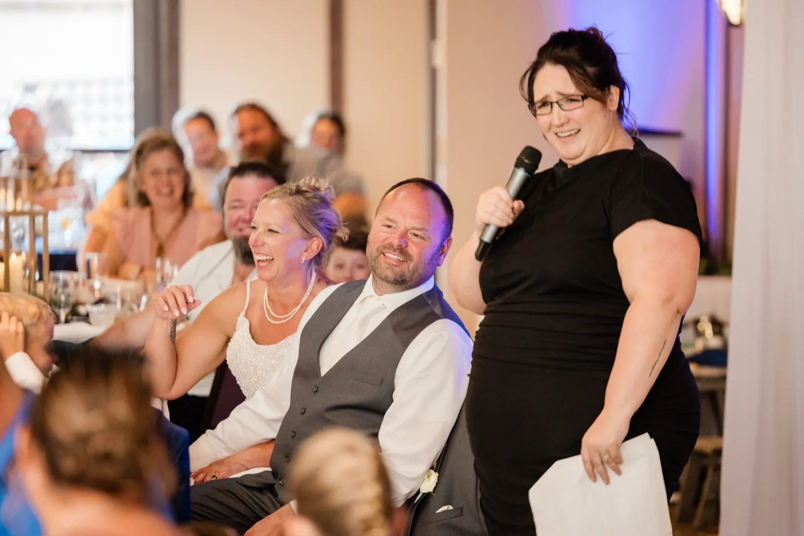 A woman in a black dress holds a microphone and notes while giving a toast as the bride and groom laugh in their seats behind her — Tim Larsen Photography, Brainerd Lakes MN