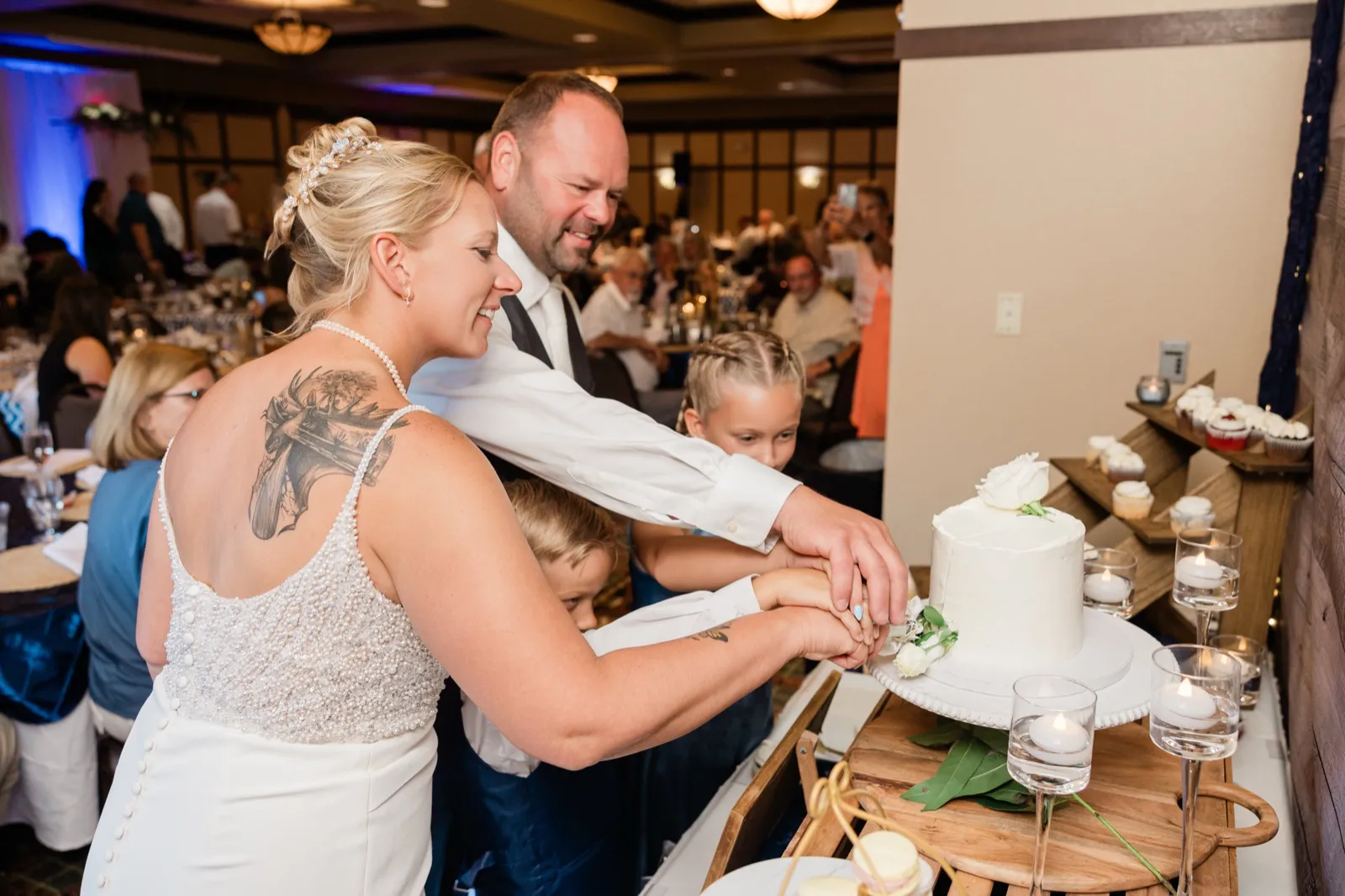 The bride and groom cut a small white wedding cake while two children reach in to help hold the knife, with guests seated at tables behind them — Tim Larsen Photography, Brainerd Lakes MN