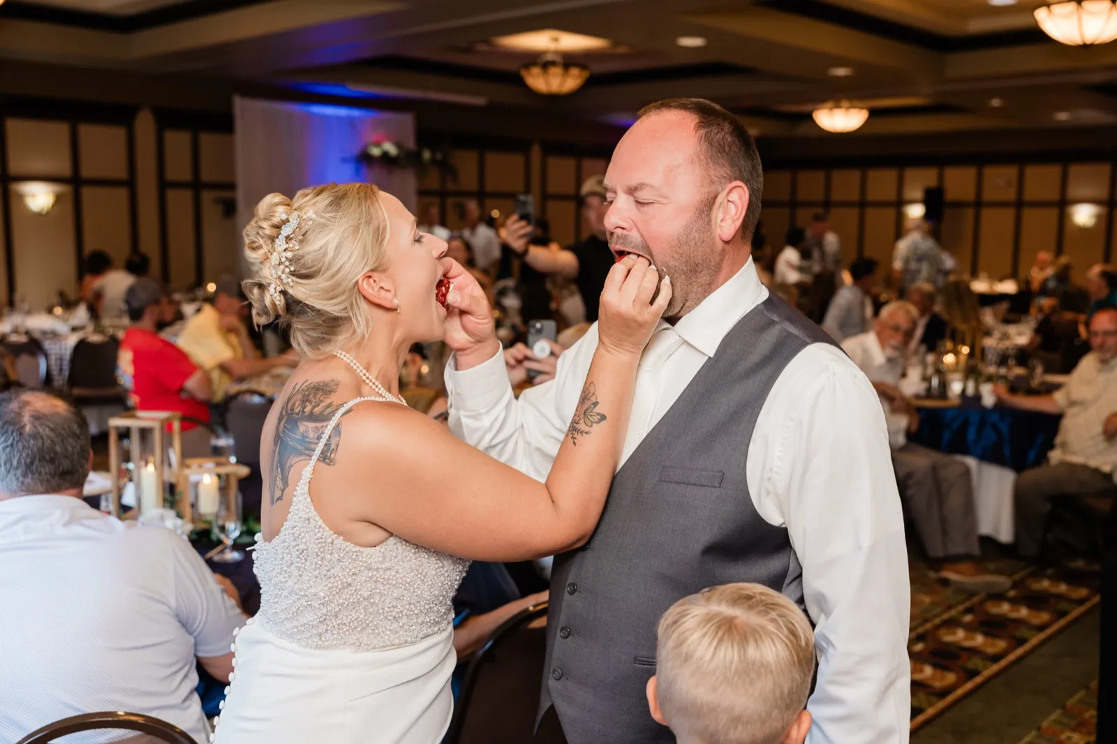The bride and groom feed each other bites of cake mouth-to-hand while guests watch from tables in a resort ballroom — Tim Larsen Photography, Brainerd Lakes MN